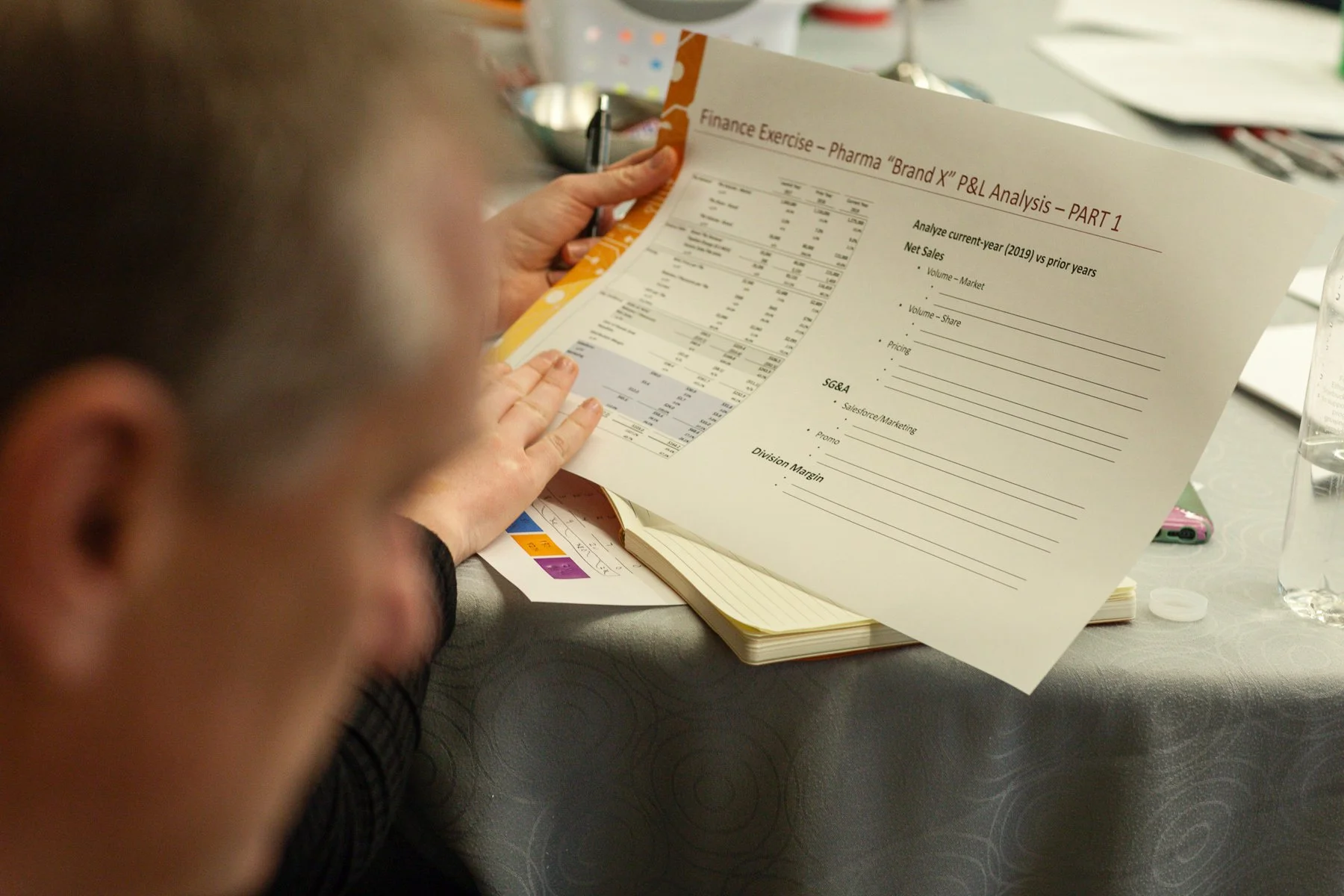 Person holding a financial analysis report titled 'Finance Exercise - Pharma Brand X P&L Analysis - PART 1' at a table with various documents, a notebook, and a glass of water nearby.