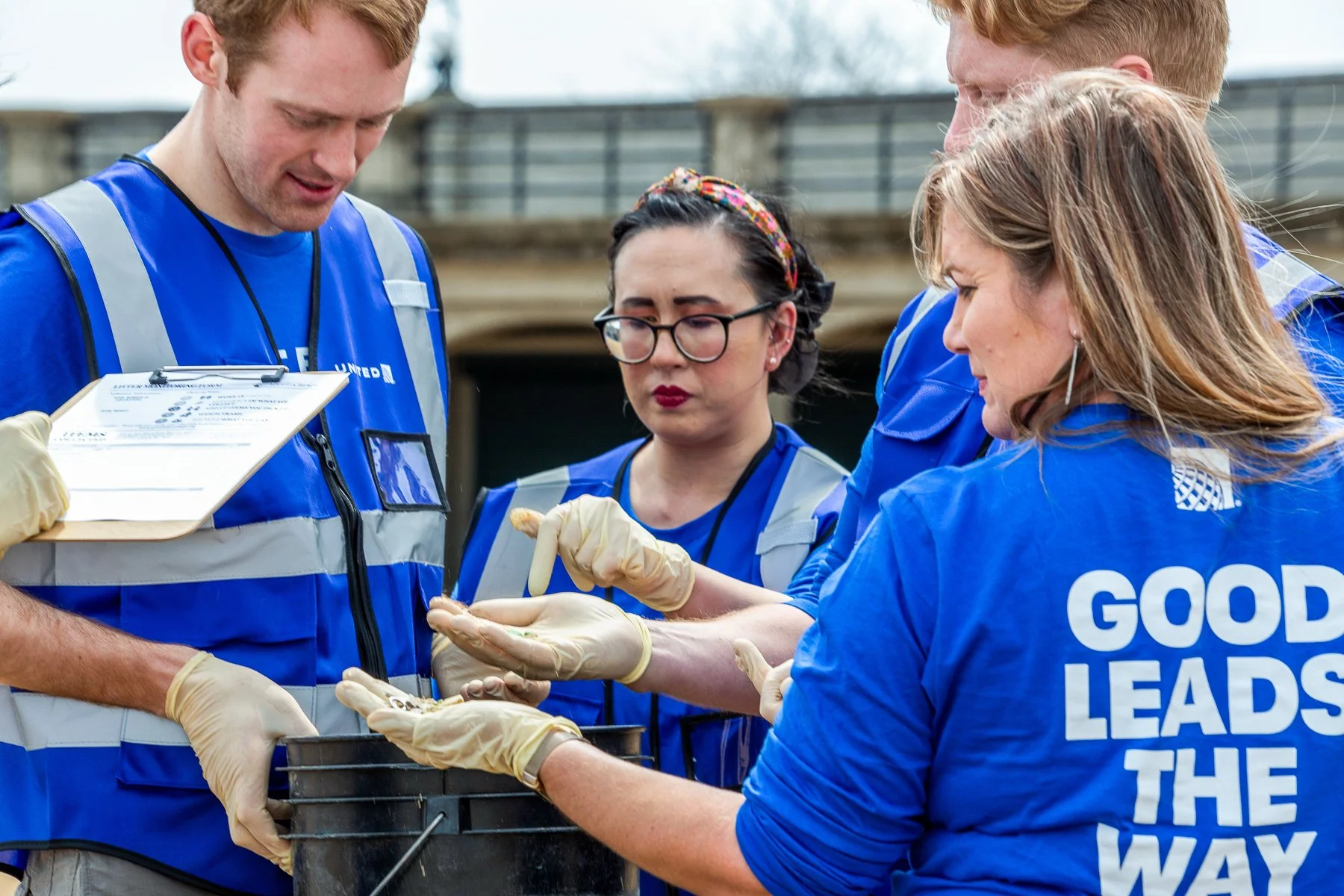 A group of people wearing blue volunteer shirts and latex gloves gathered around an outdoor medical setup, examining and handling a small animal or specimen during a community service event.