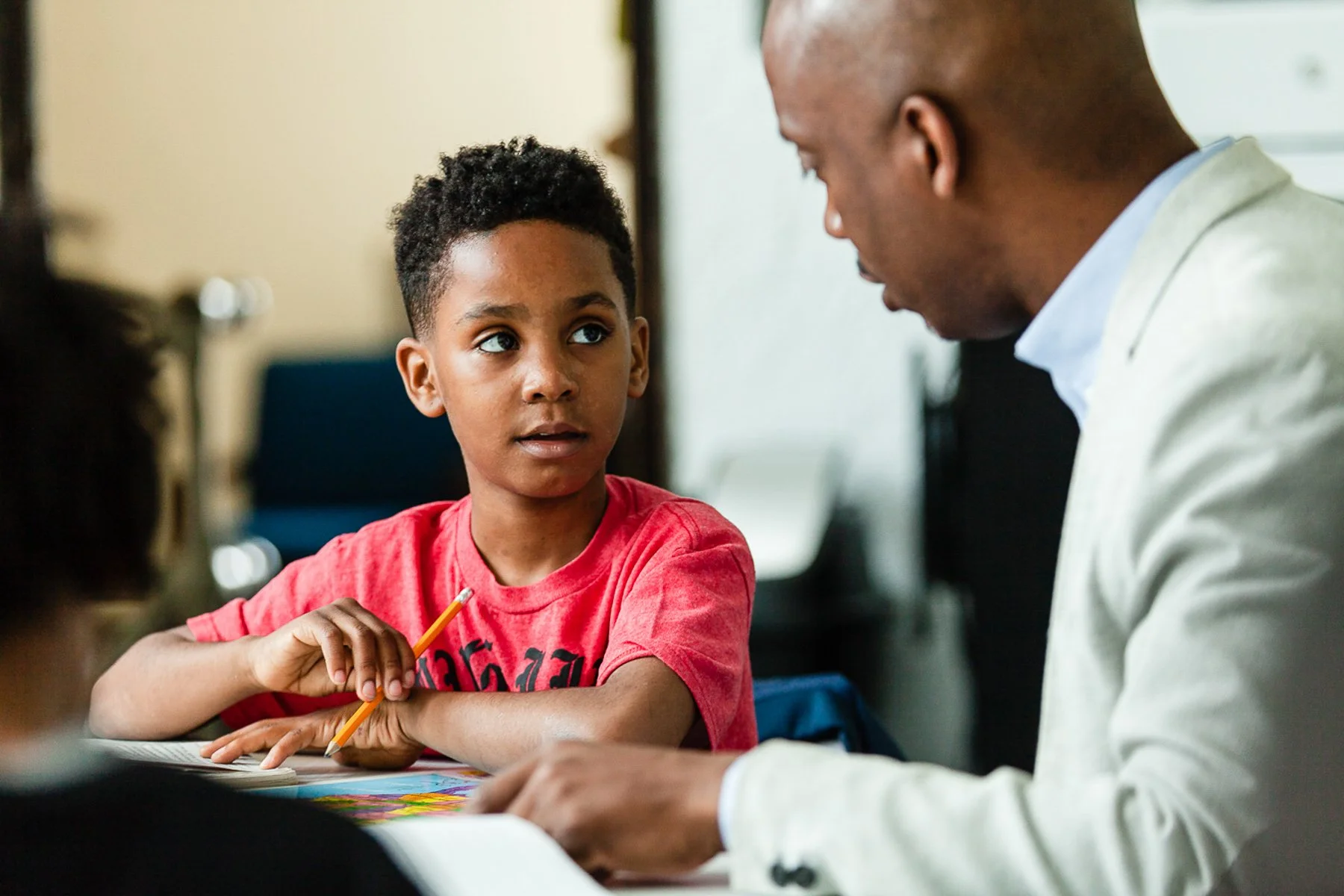 A young boy with dark curly hair and a red shirt looks attentively at an adult man with a bald head and light-colored blazer, who is reading from a book in a classroom or study setting.
