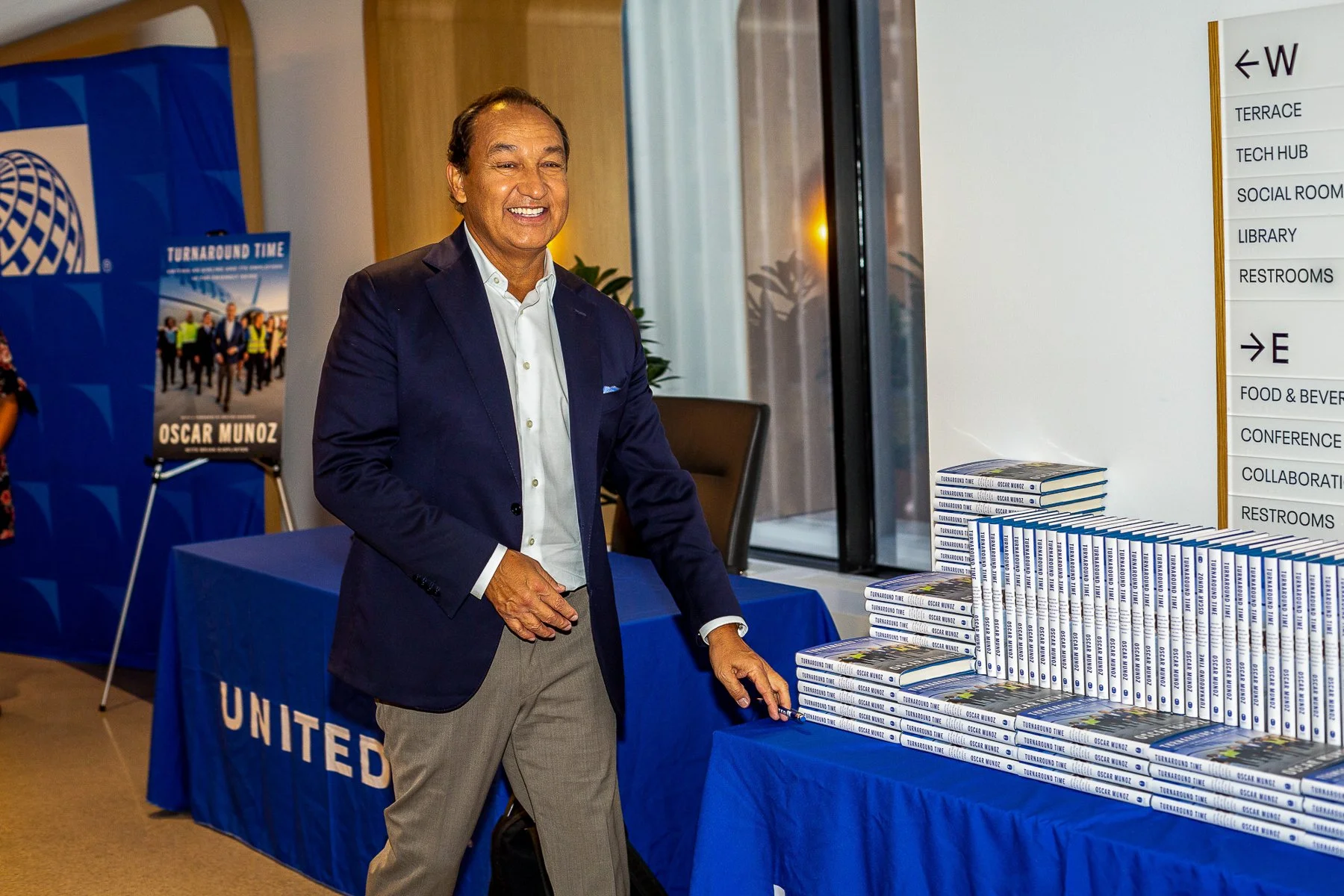A man in a navy blazer and light gray pants standing at a table with stacks of books titled 'Turnaround Time' by Oscar Munoz. Behind him is a blue United Airlines table cover and a poster of workers in safety vests. The setting appears to be a confer