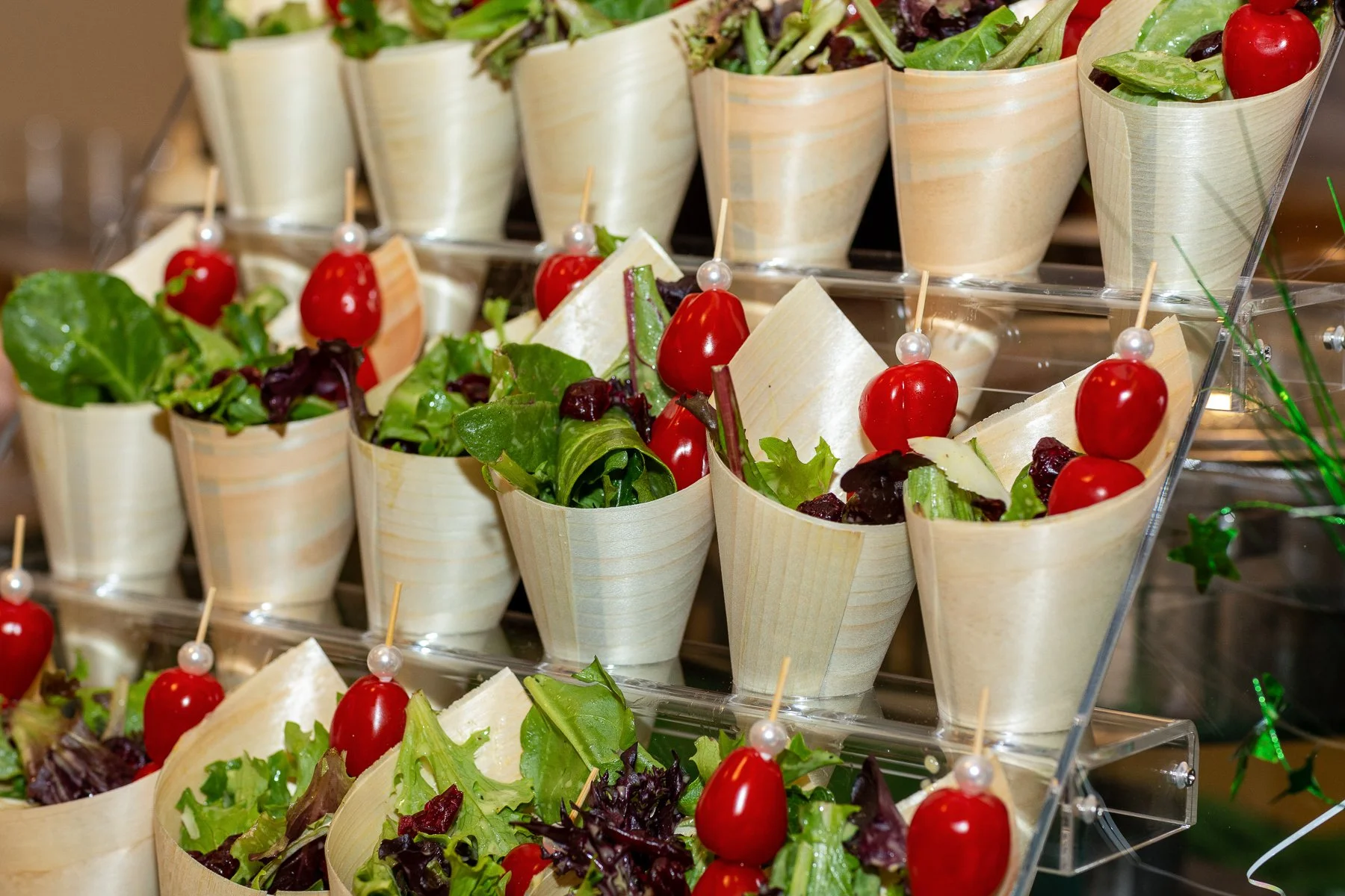 Multiple cups filled with fresh green salad, cherry tomatoes, and slices of cheese, arranged on a multi-tiered transparent display stand.