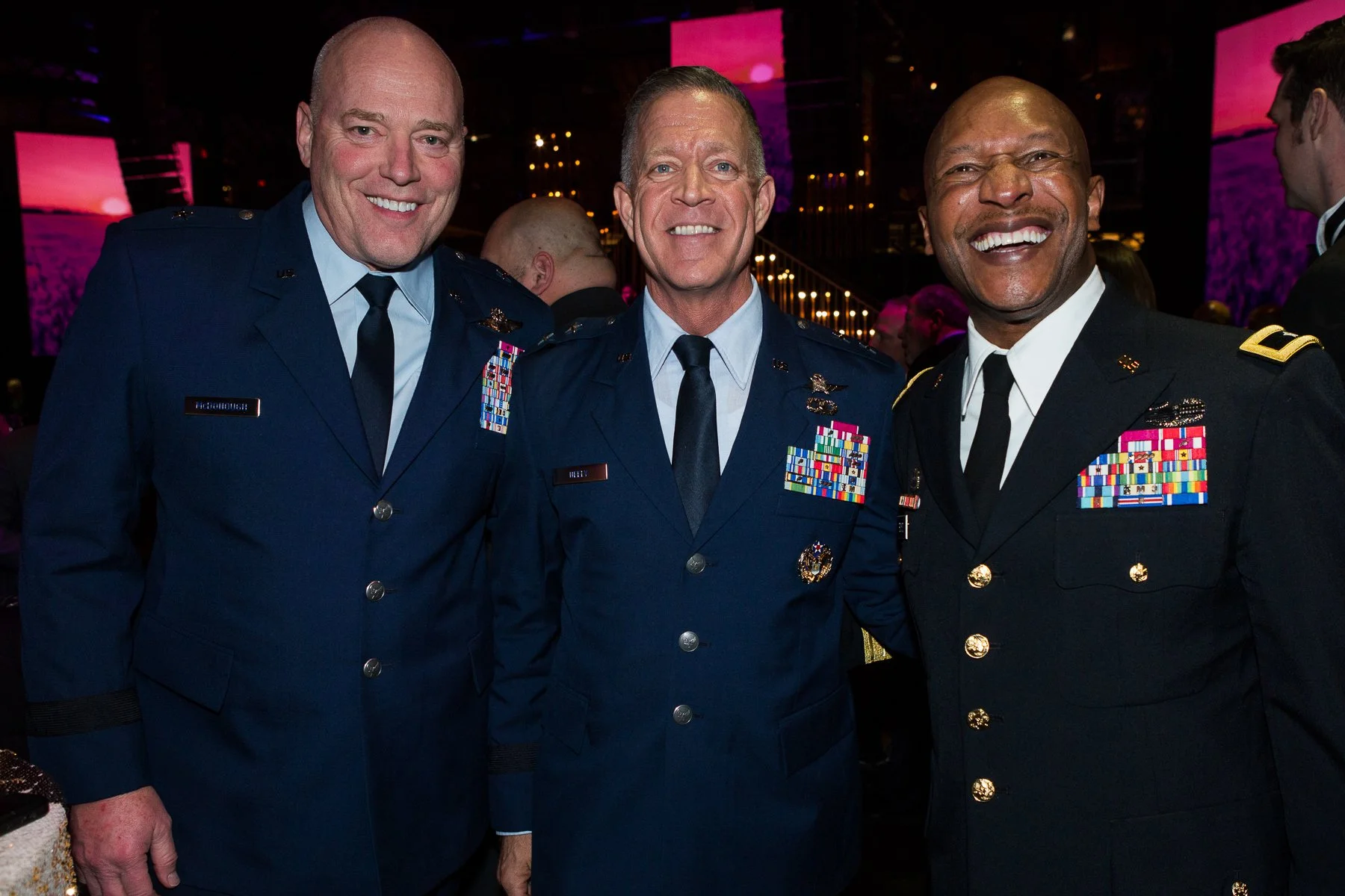 Three men in military uniforms smiling at an event, with a pink sunset background on screens behind them.