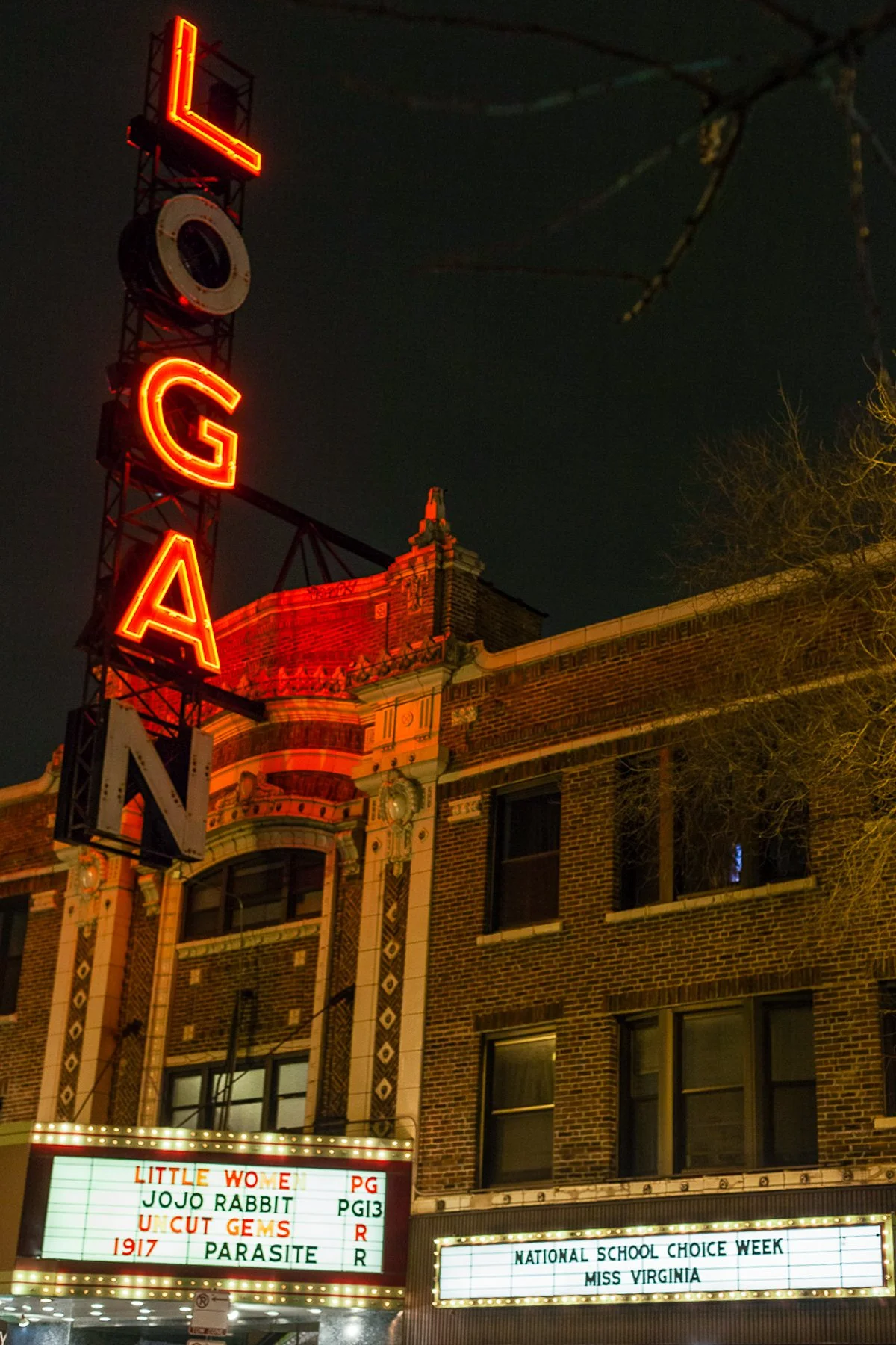Night view of a historic brick theater with a neon sign that reads 'LOEGA' and an illuminated marquee displaying movie titles and event information, including 'NATIONAL SCHOOL CHOICE WEEK' and 'MISS VIRGINIA.'