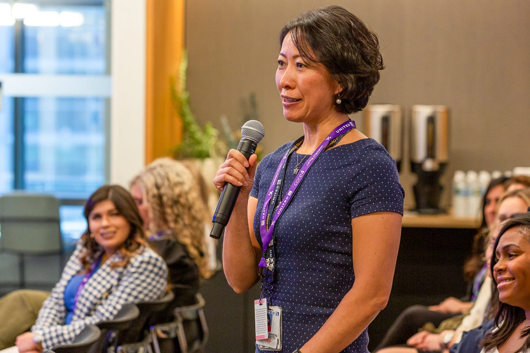 A woman with short dark hair, wearing  blue dress and a lanyard, is speaking into a microphone at a conference or seminar. She is surrounded by an audience, including women of diverse backgrounds, seated and attentively listening.©2026 Steven Barston