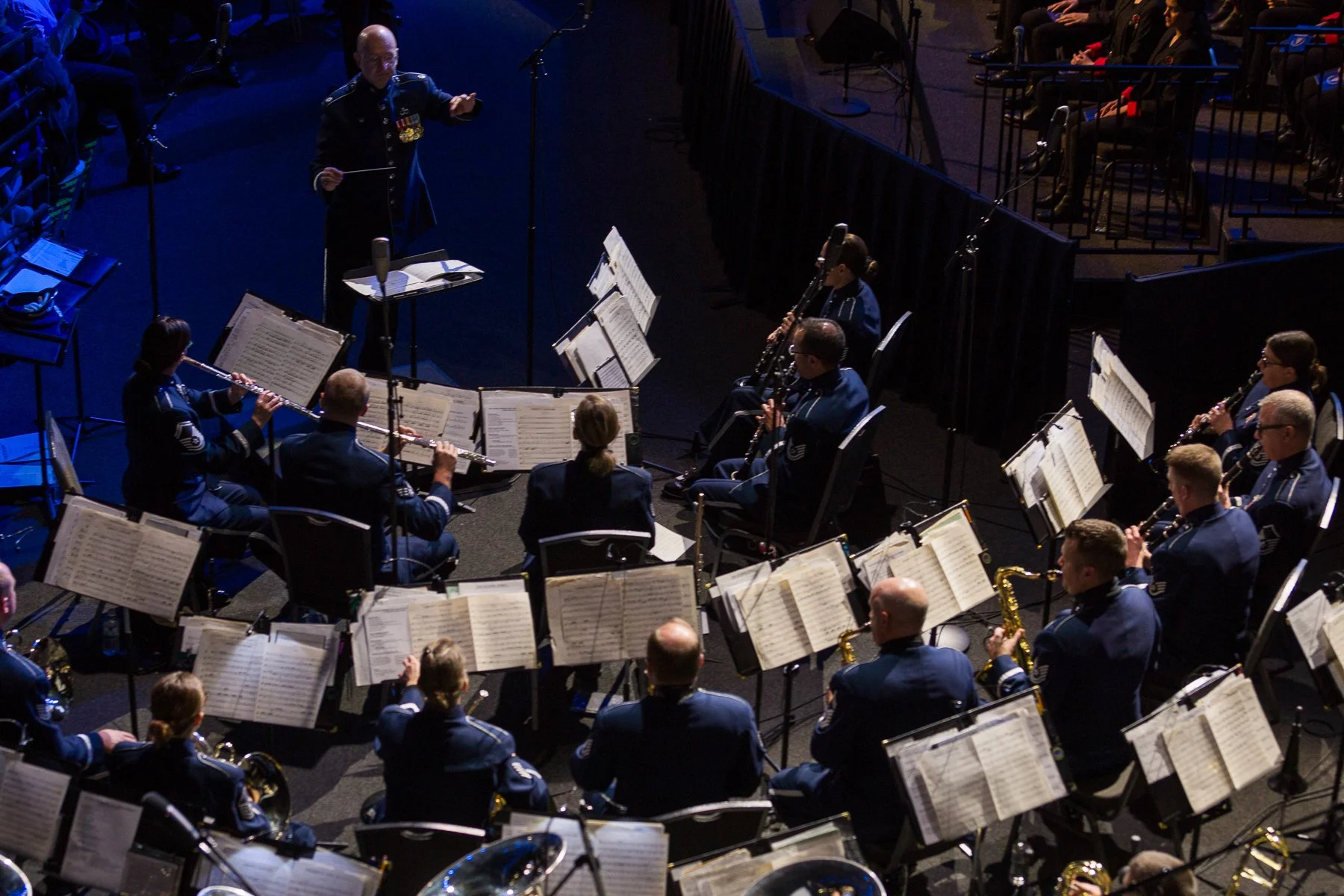 A military band performing on stage with musicians in uniform playing various wind instruments, directed by a conductor standing in front of them.