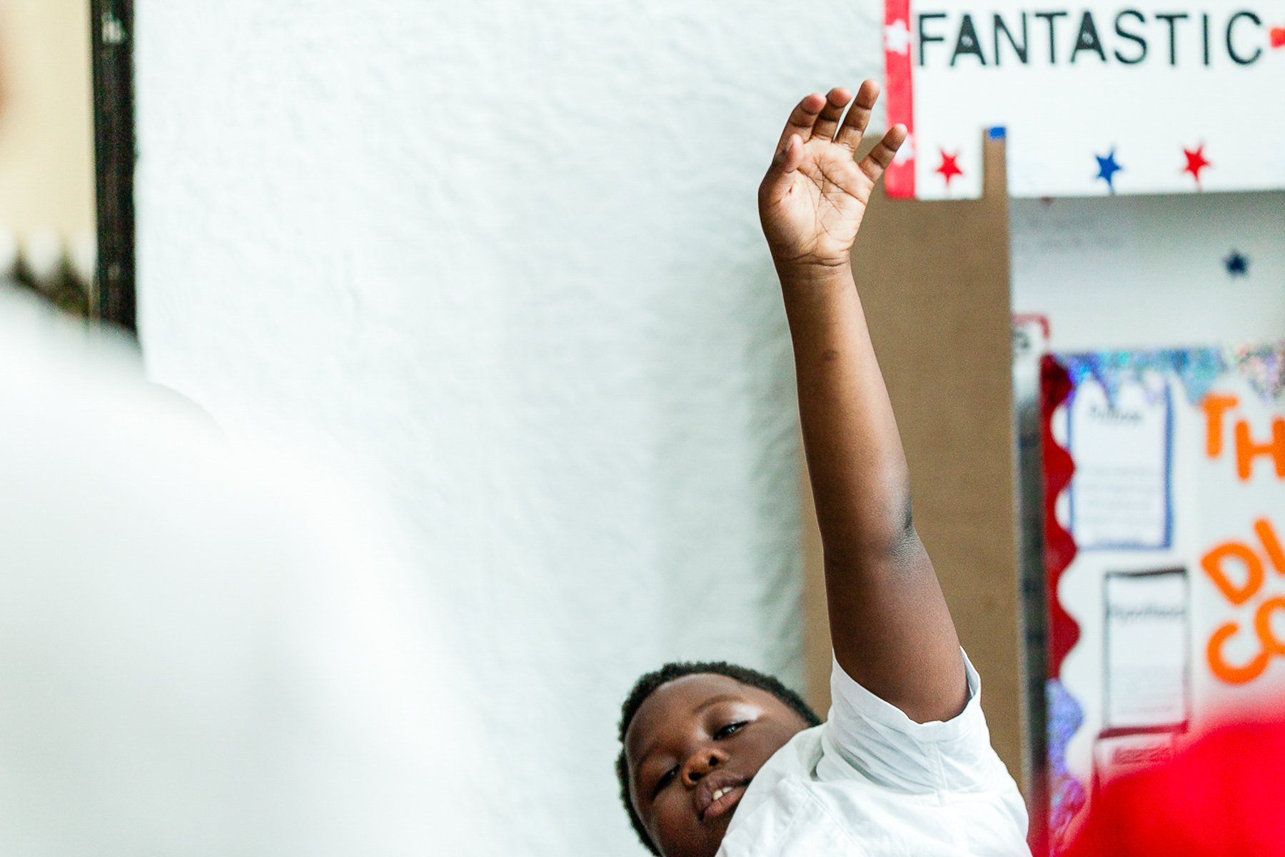 Young student raising hand in classroom with educational posters on the wall.