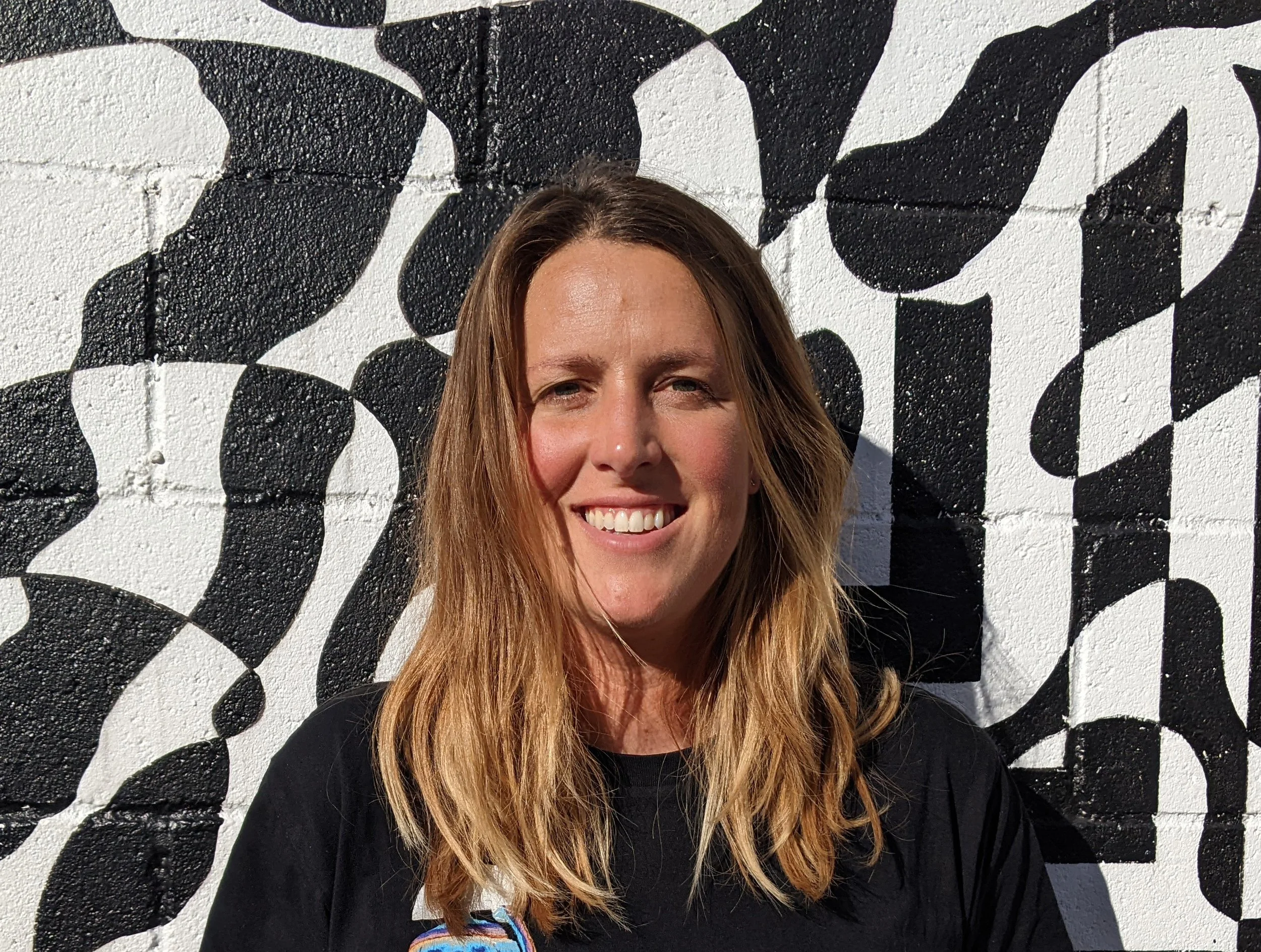 A woman with long, wavy blonde hair smiling and showing teeth, standing in front of a black and white painted wall with large, abstract patterns.