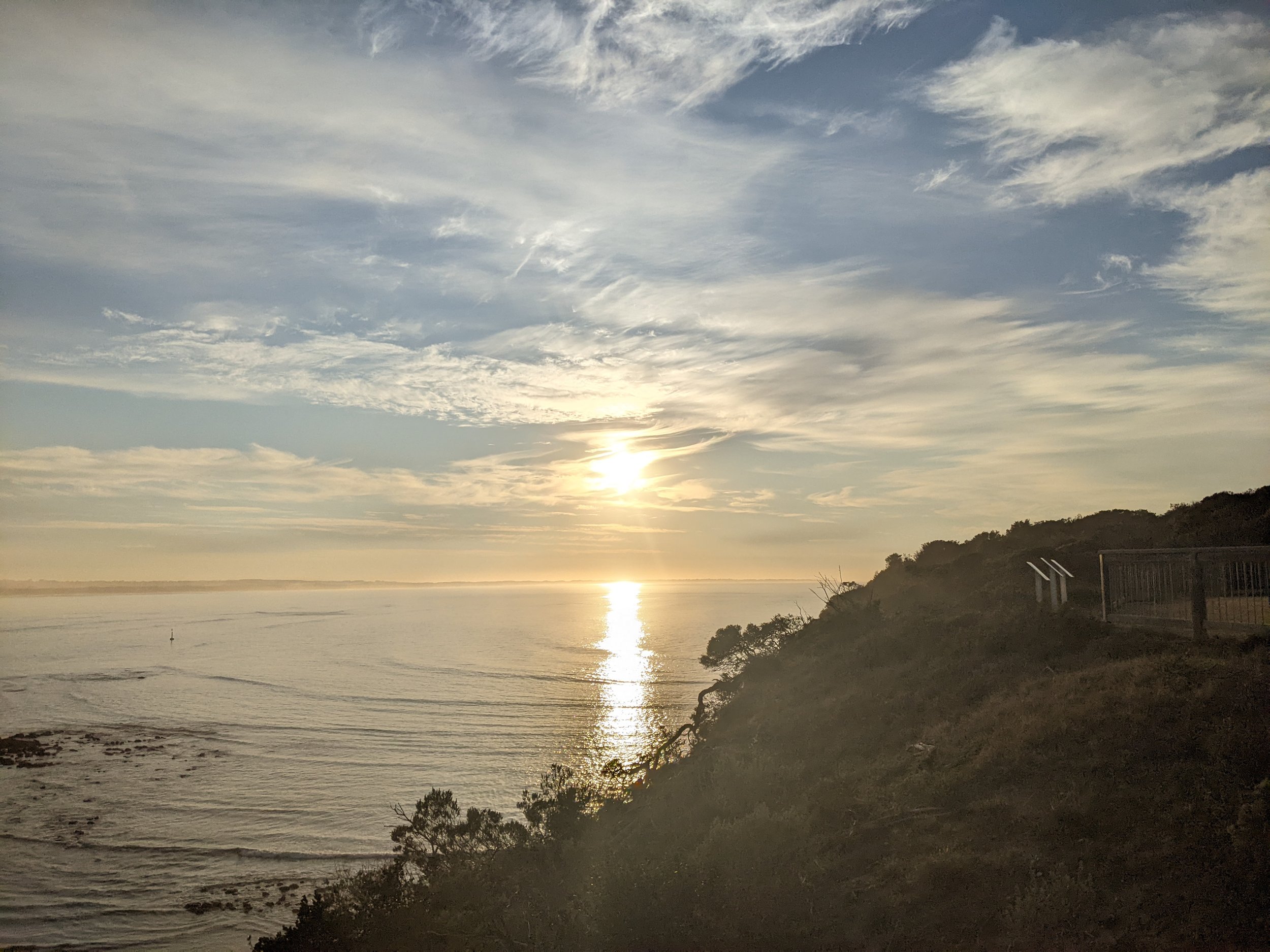 Sun setting over a calm ocean with a partly cloudy sky and silhouetted trees on the shoreline. Photo taken from the Bluff at Barwon Heads.