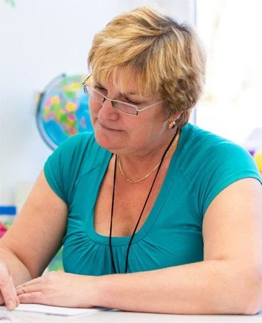 A woman with short blonde hair and glasses sitting at a desk, writing or working, with a colorful world globe in the background.