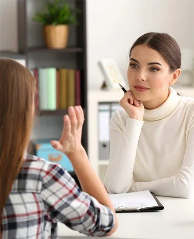 A woman in a white turtleneck listening to a young girl in a checkered shirt during a conversation in an office setting.