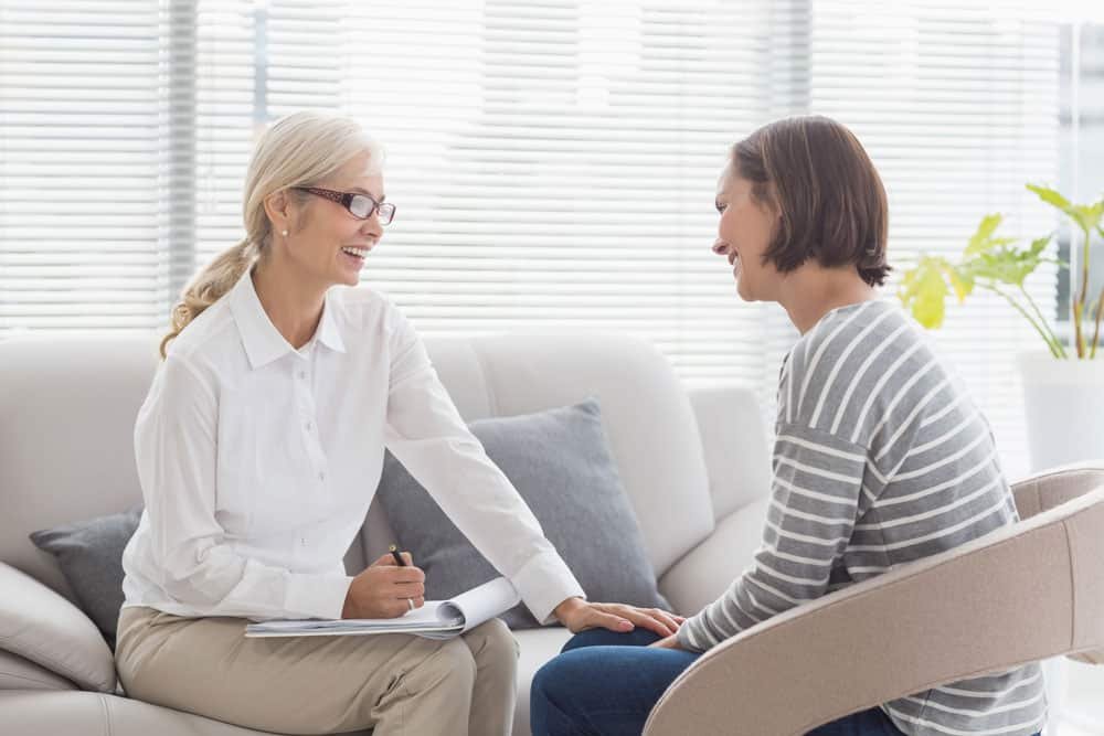 A woman in a striped shirt talking to a professional counselor or therapist with glasses, in a bright, modern office with blinds and plants.