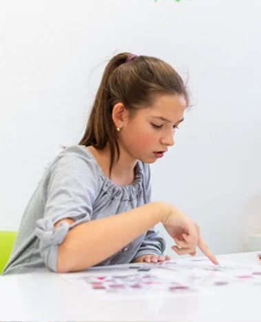 A young girl with brown hair tied in a ponytail sitting at a table, pointing at something on the table, in a well-lit room with a white wall.