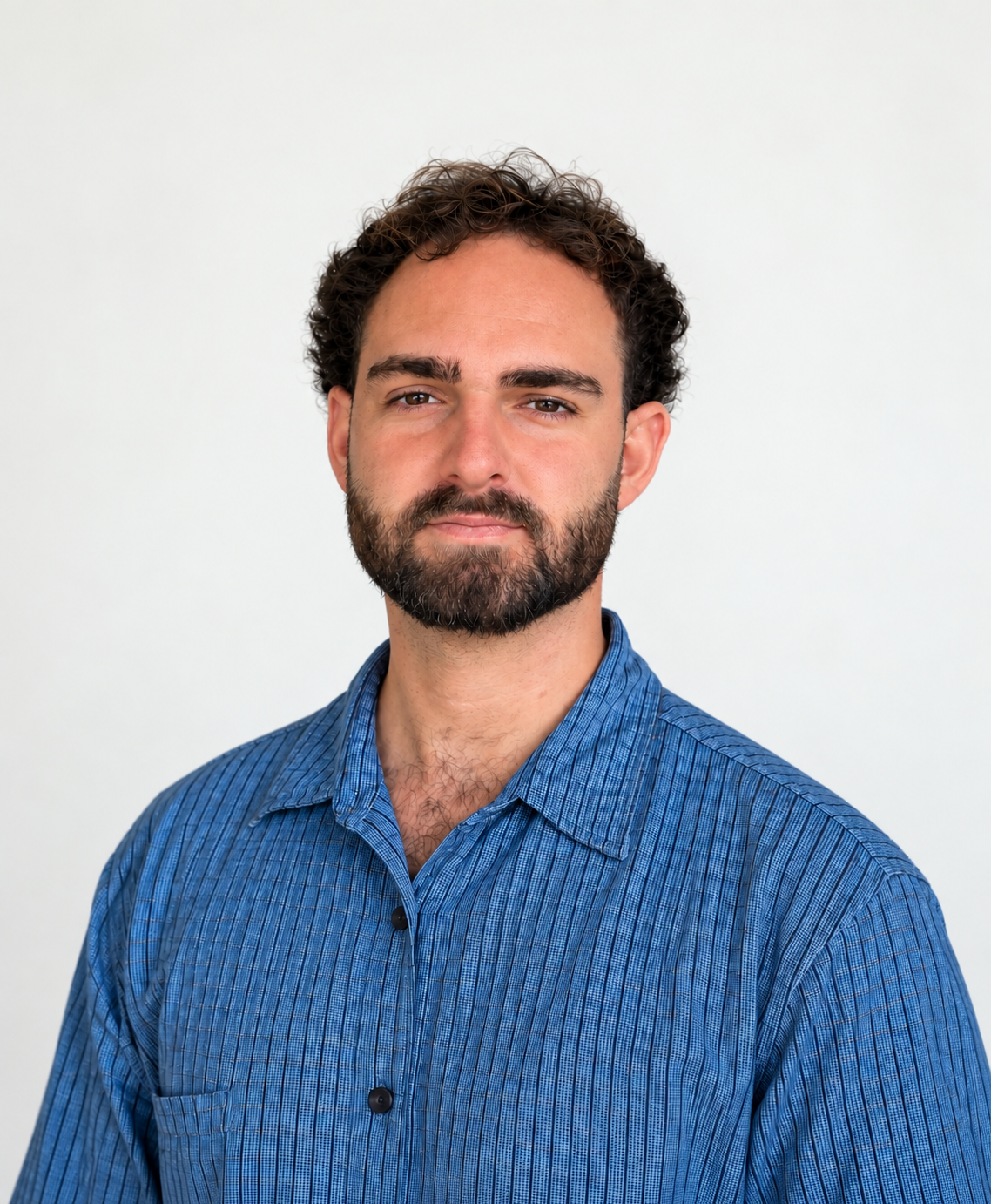 Portrait of a man with dark, curly hair and a beard, wearing a blue striped button-up shirt, standing against a plain white background.