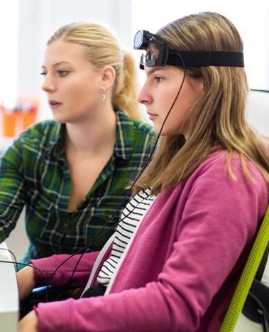 Two women seated at a table, one wearing a headlamp with a magnifying glass, examining a device, suggesting a scientific or medical setting.