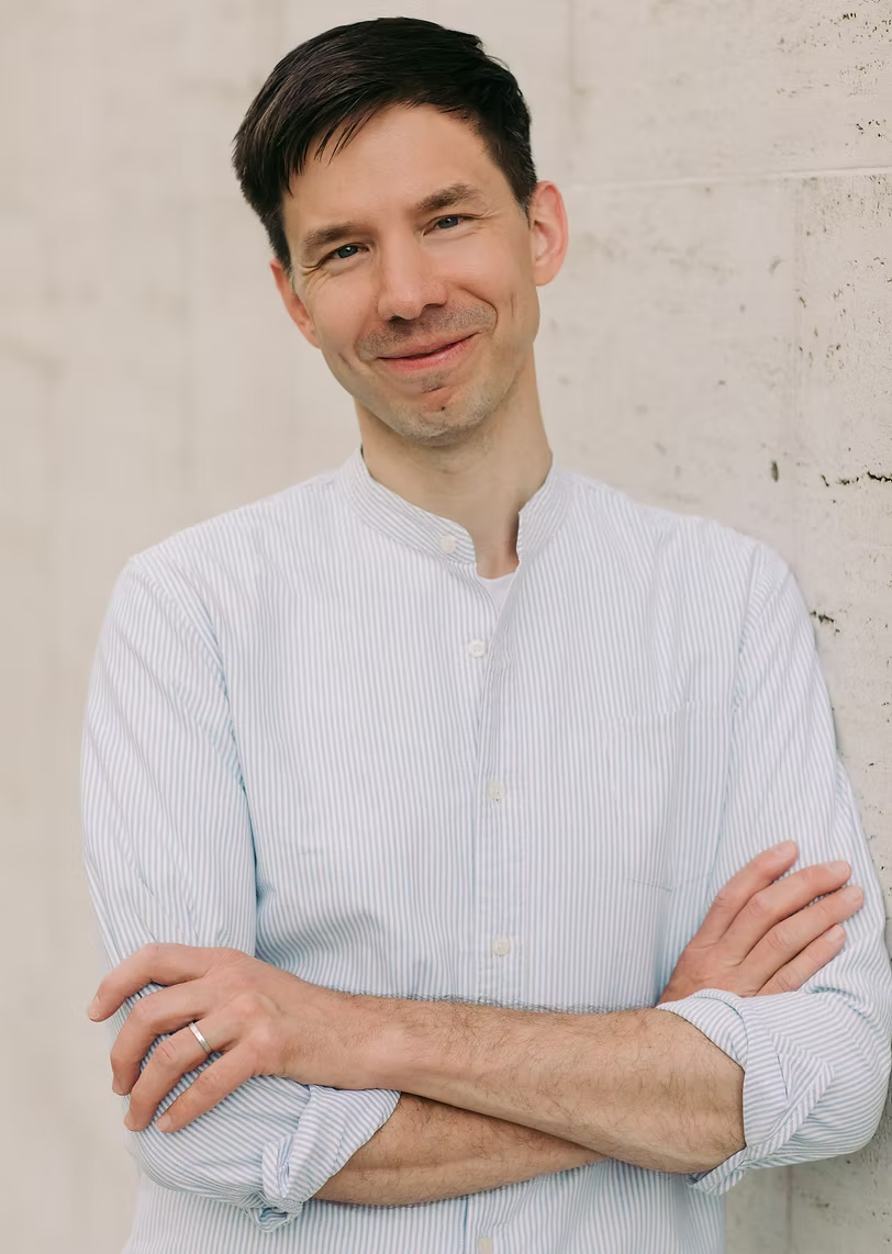 A man with dark hair and a light shirt leaning against a white brick wall, smiling with arms crossed.