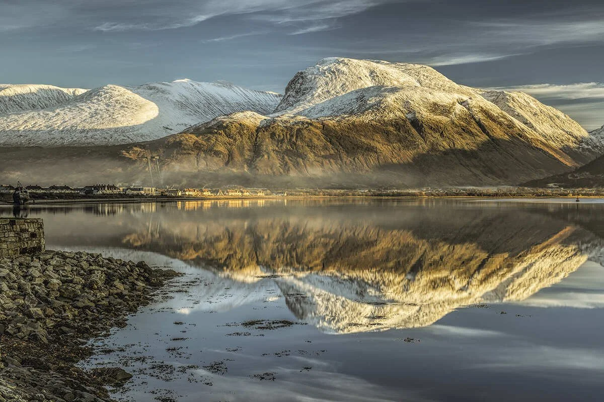 Snow-capped mountains reflected in a calm body of water with a rocky shoreline in the foreground.