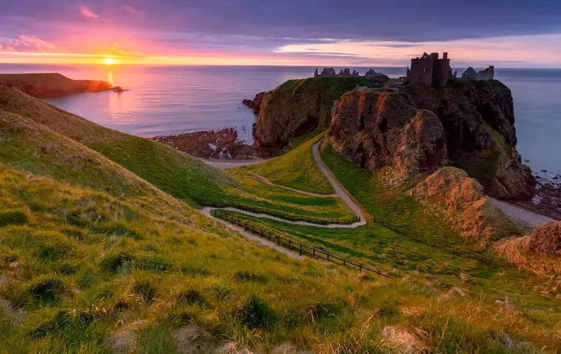 Scenic view of a coastline with a castle on a cliff, grassy hills in the foreground, and a sunset over the ocean.