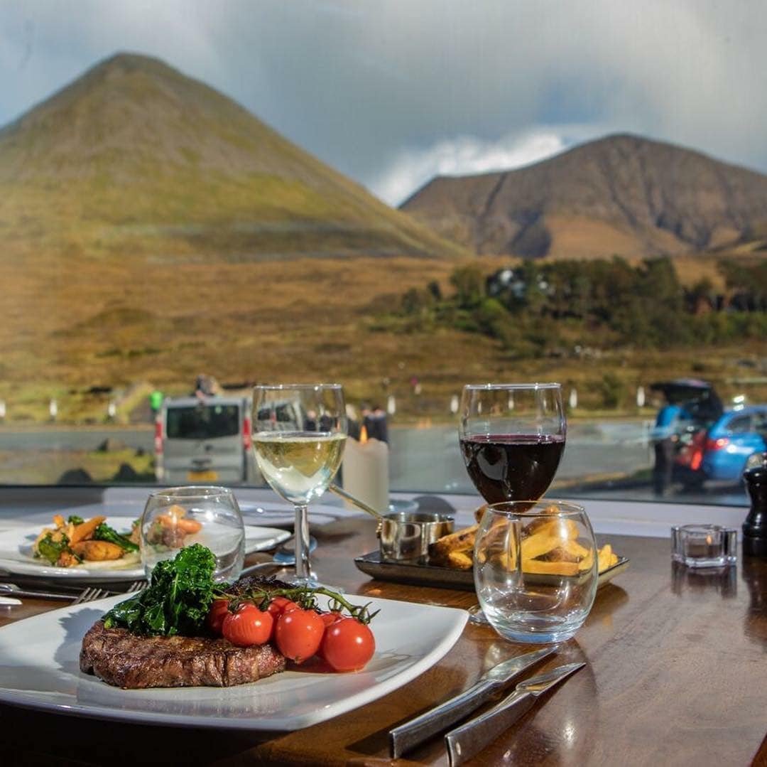 Dinner table with steak, cherry tomatoes, vegetables, wine glasses with white and red wine, and a view of mountains outside the window.