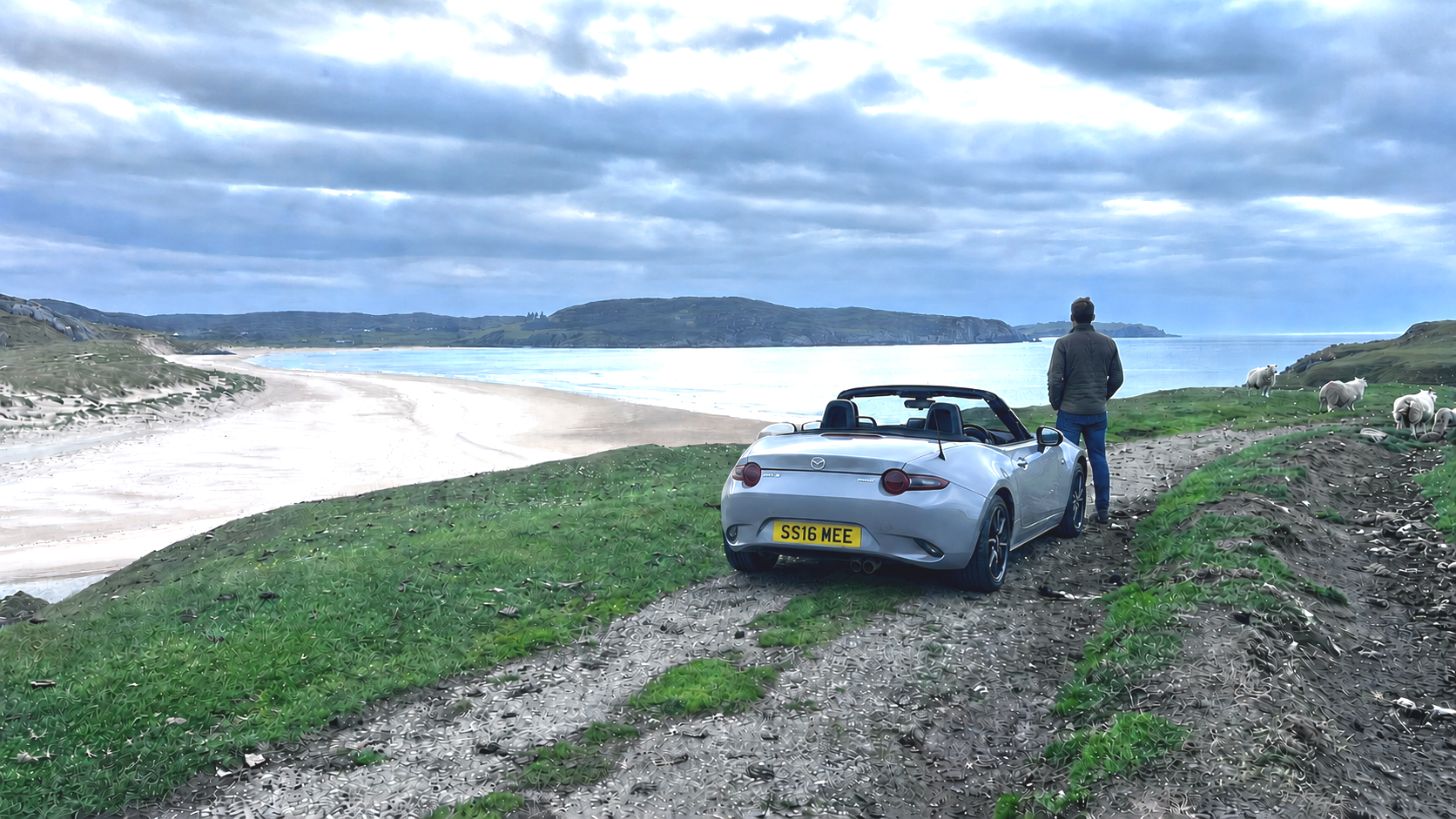 Man standing near a silver Miata convertible sports car on a grassy path overlooking a beach and ocean, with sheep grazing nearby and hills in the distance under a cloudy sky.