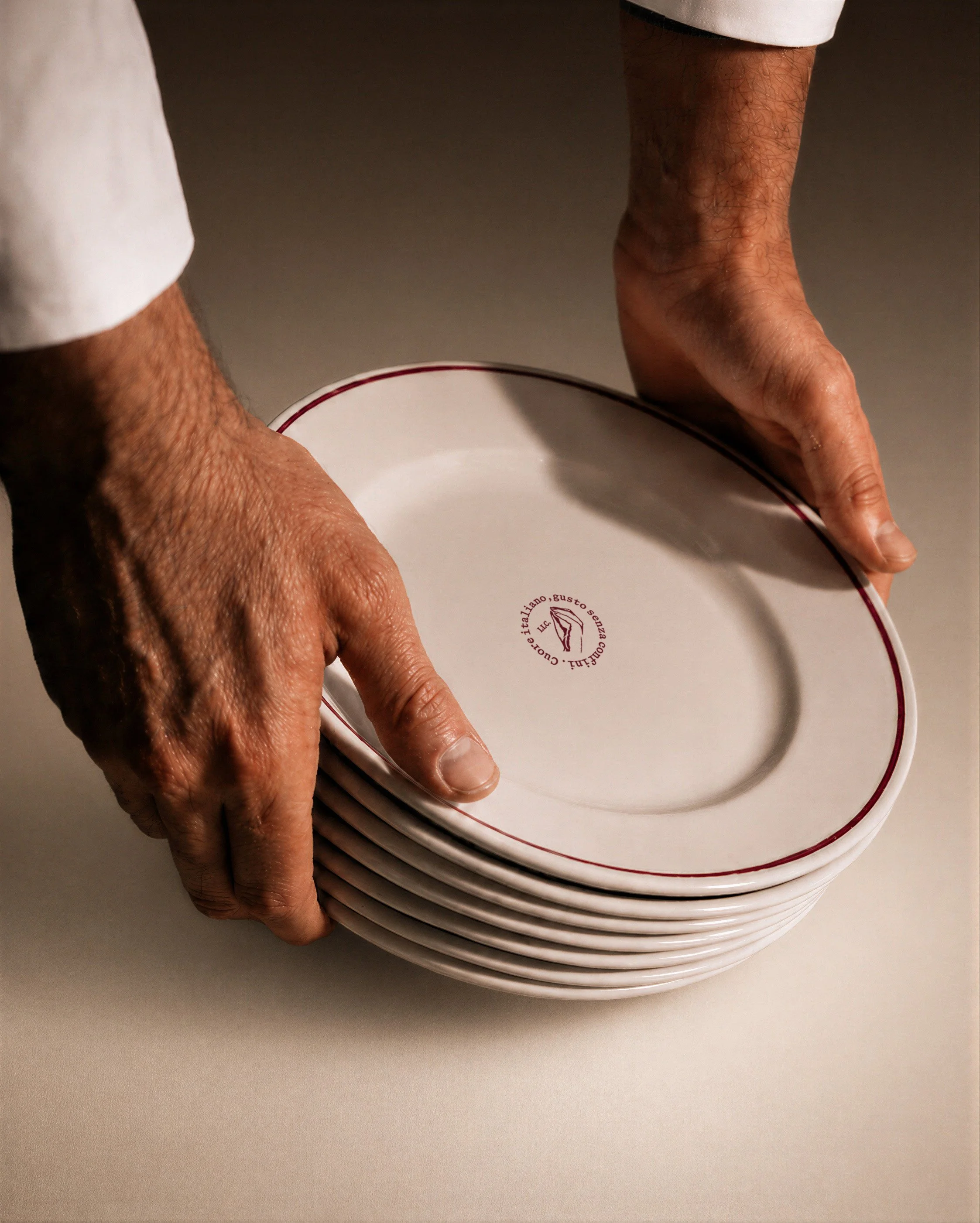 Multiple white ceramic plates stacked together on a plain light-colored surface, with part of a person's hand and arm visible as they hold the plates.