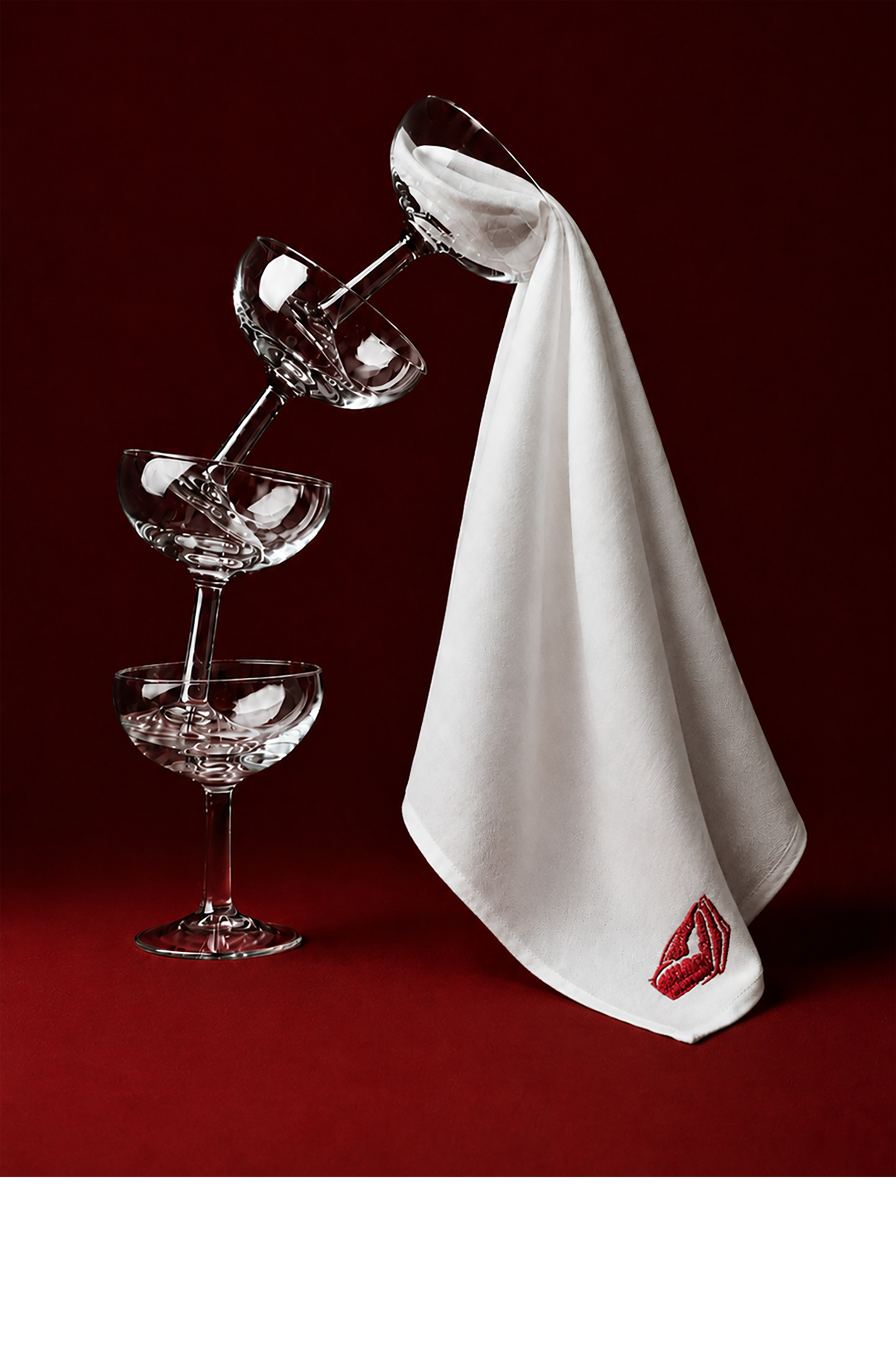 Three empty Martini glasses balanced on top of each other with a white napkin hanging from the top glass, embroidered with a red kiss mark, against a dark red background.