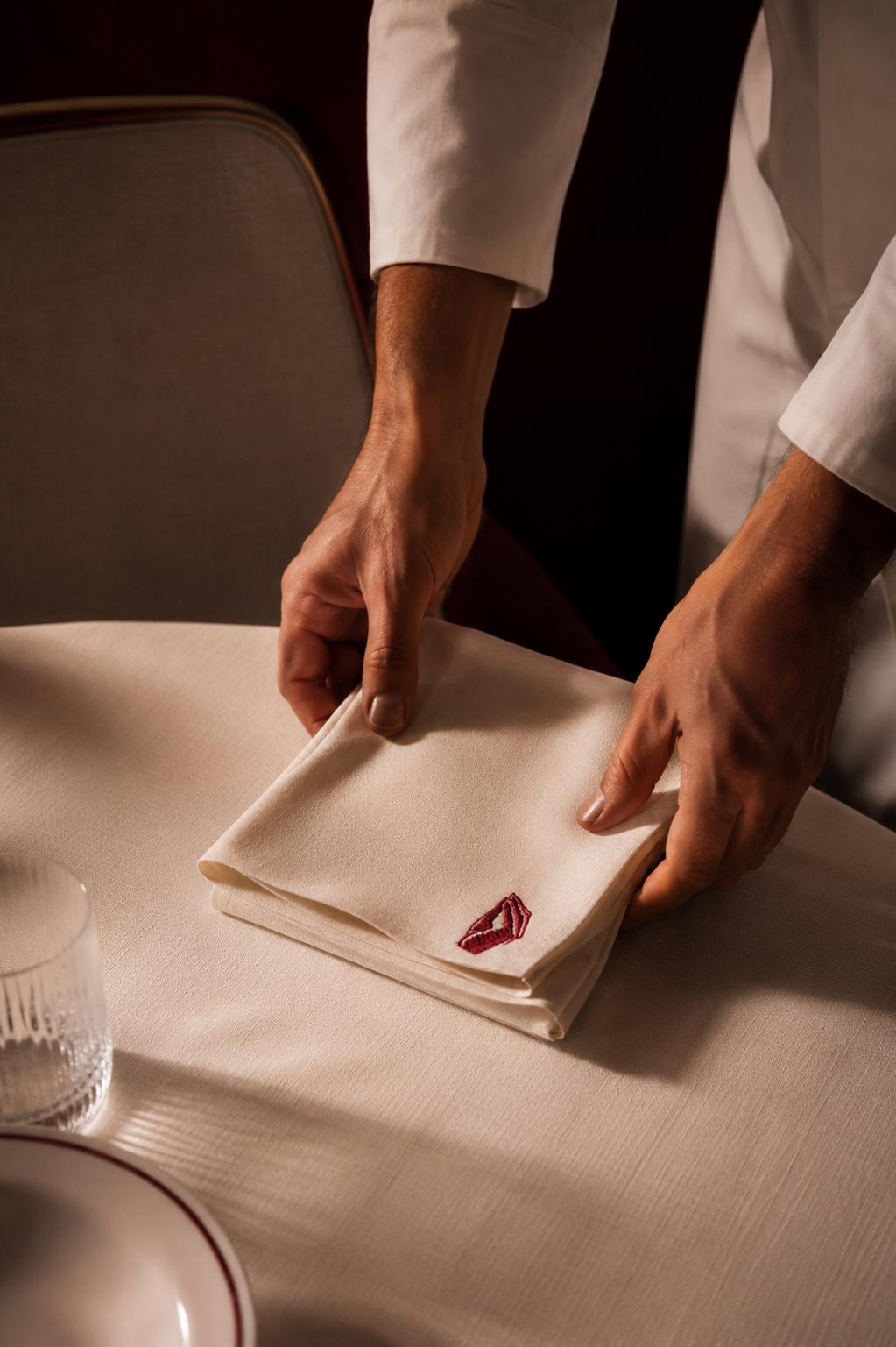 A person in a white shirt is folding a white napkin with a red embroidered lipstick mark on it at a dining table.