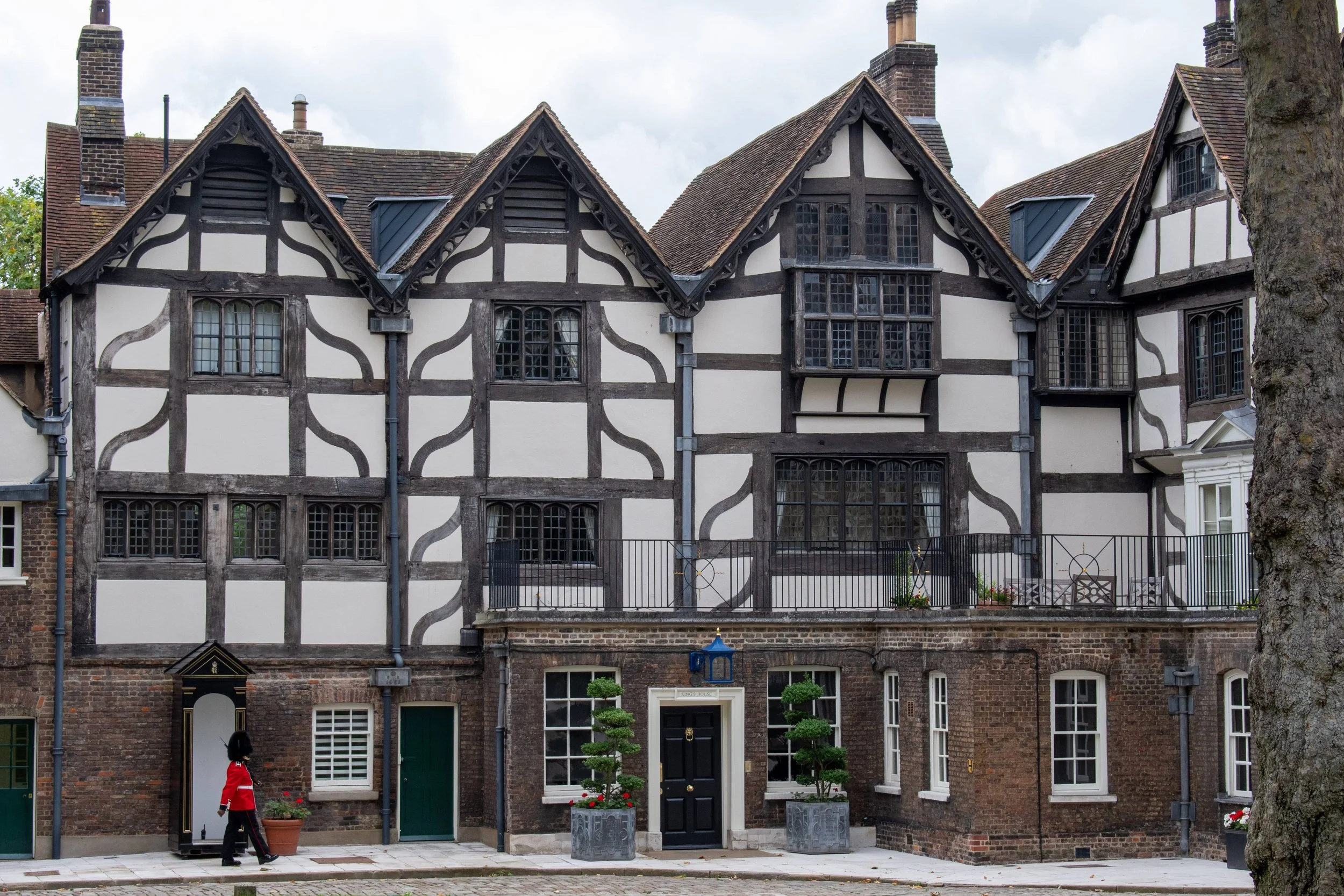 A row of old, historic Tudor-style houses with black timber framing and white walls, brick bases, pitched roofs with chimneys, and variously shaped windows. A person dressed in a red jacket and black pants is walking in front of the buildings, and there are potted plants and small trees near the doors.