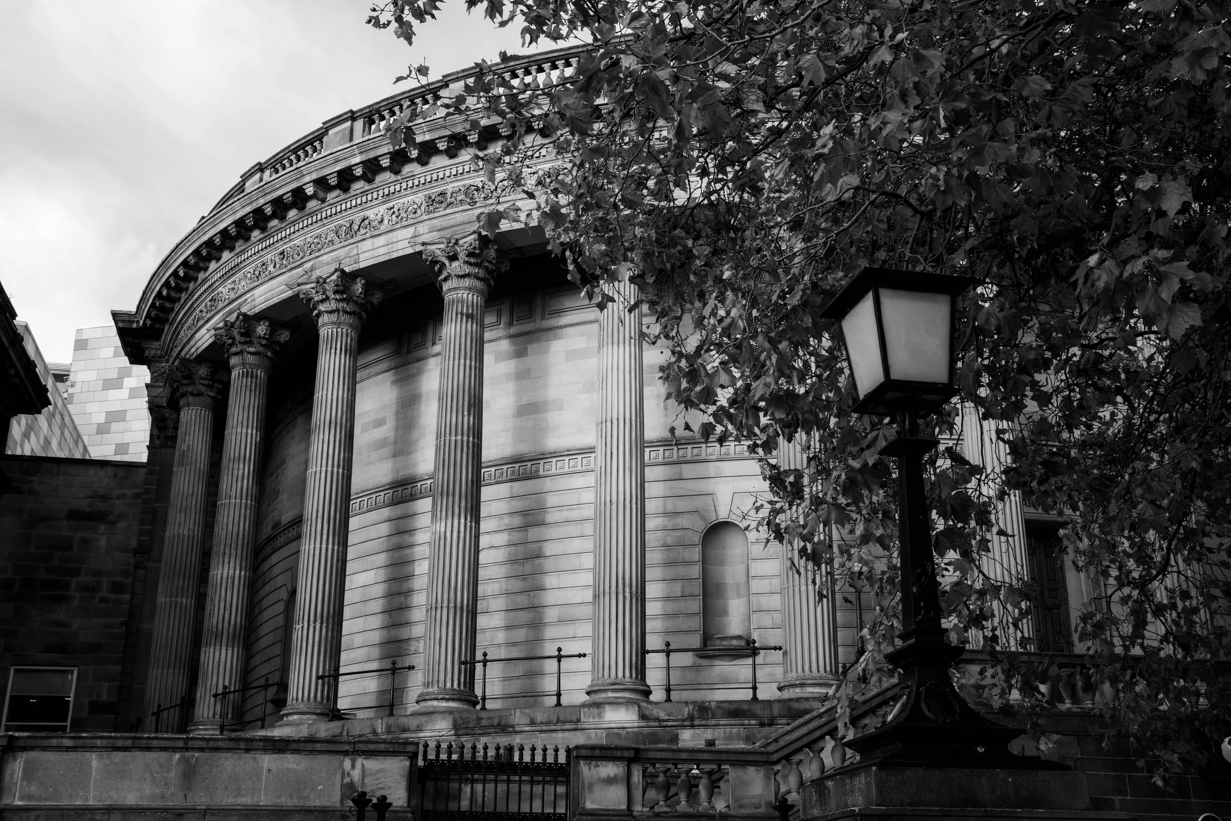 A black and white photograph of a historic building with classical architecture, featuring tall columns with ornate capitals and decorative friezes. There are leafy trees partially obscuring the building, and a street lamp is visible in the foreground.