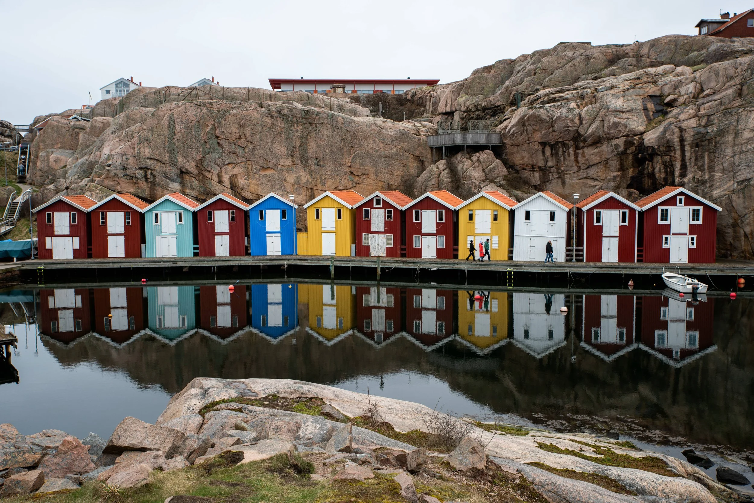 Colorful small houses with red, yellow, blue, and white exteriors are lined along the waterfront, reflected in the calm water. Large rocks and a grassy area are in the foreground, with a rocky hill and some buildings on top in the background.