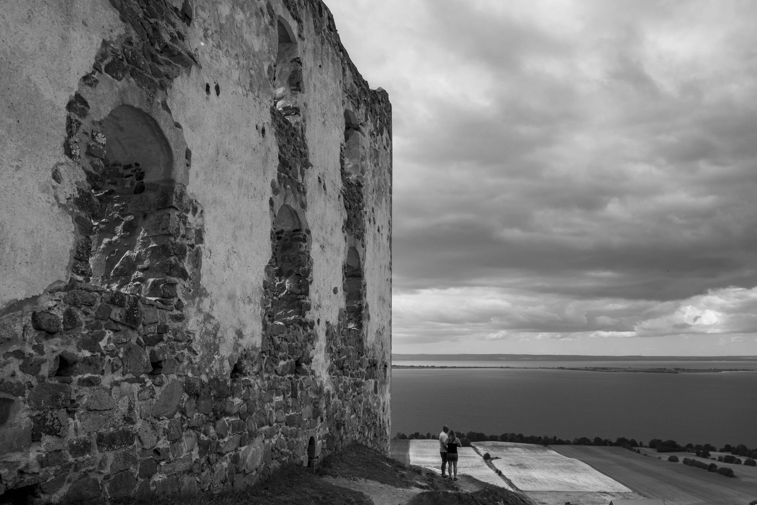 A black and white photo of an old stone building near a large body of water under a cloudy sky, with two people holding hands walking on a road beside the building.