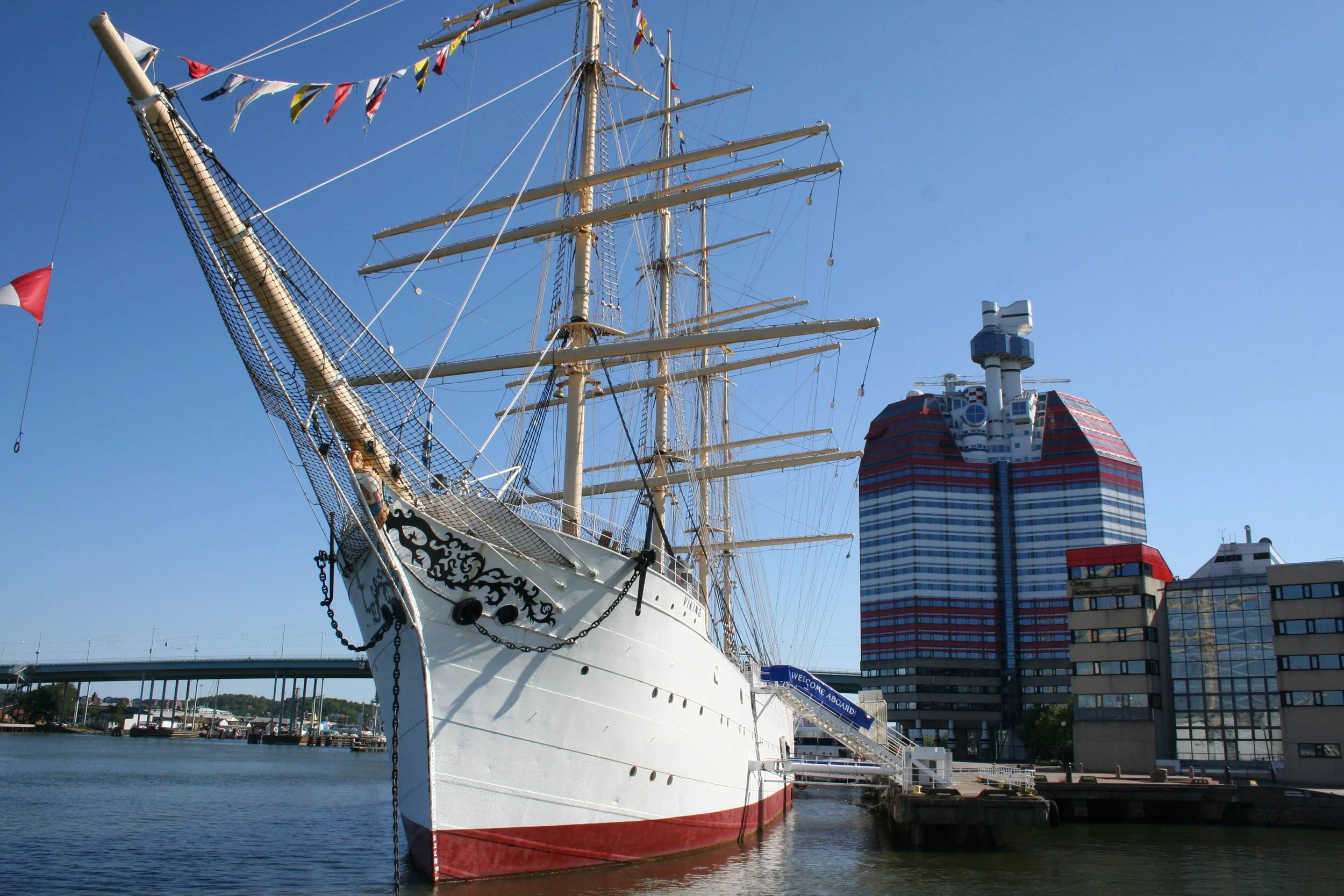 A large white sailing ship docked at a pier, with tall masts and colorful flags, in an urban harbor with modern buildings in the background.