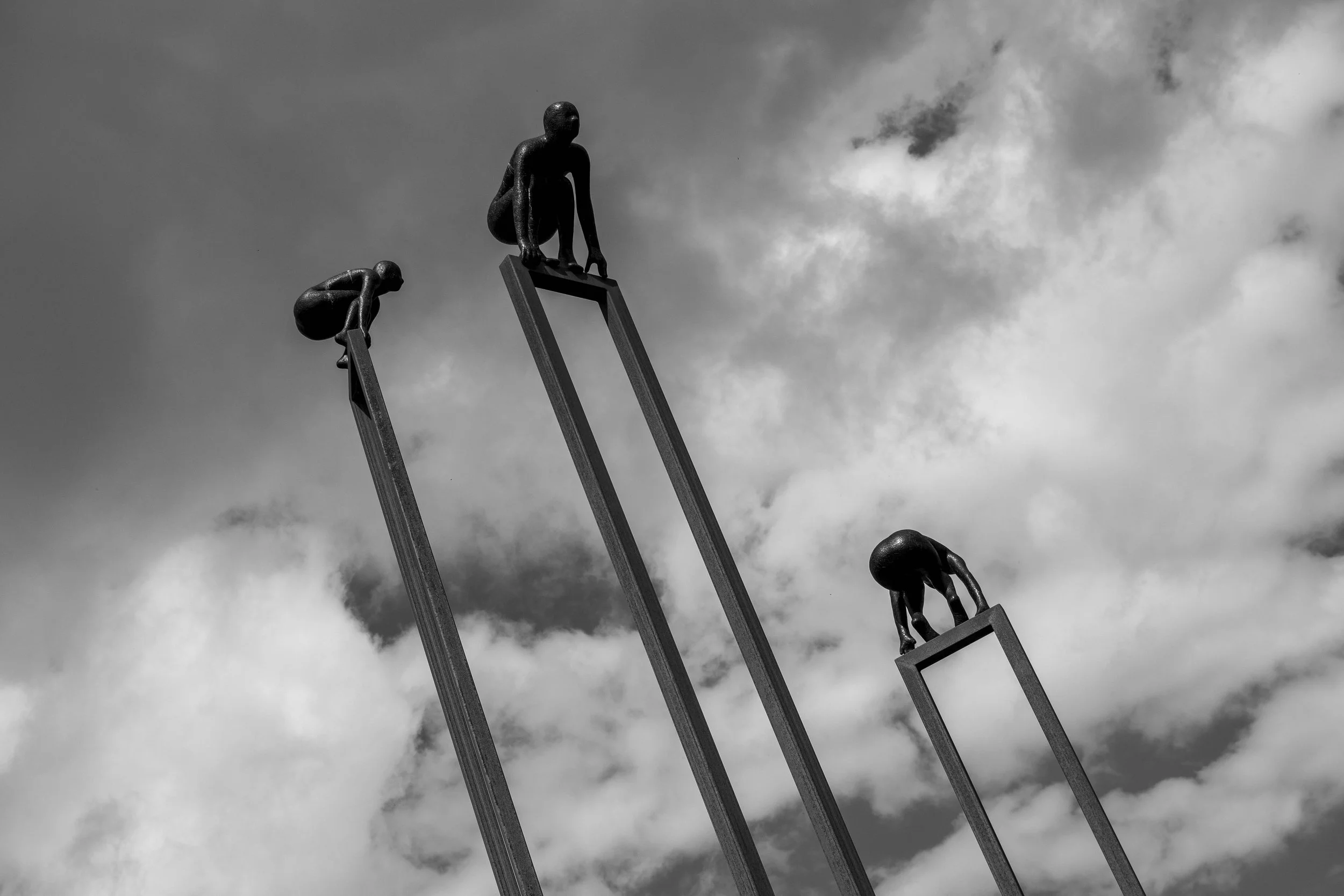 Black and white photo of small human-like figures climbing tall, thin ladders against a cloudy sky, with one figure at the top reaching out.