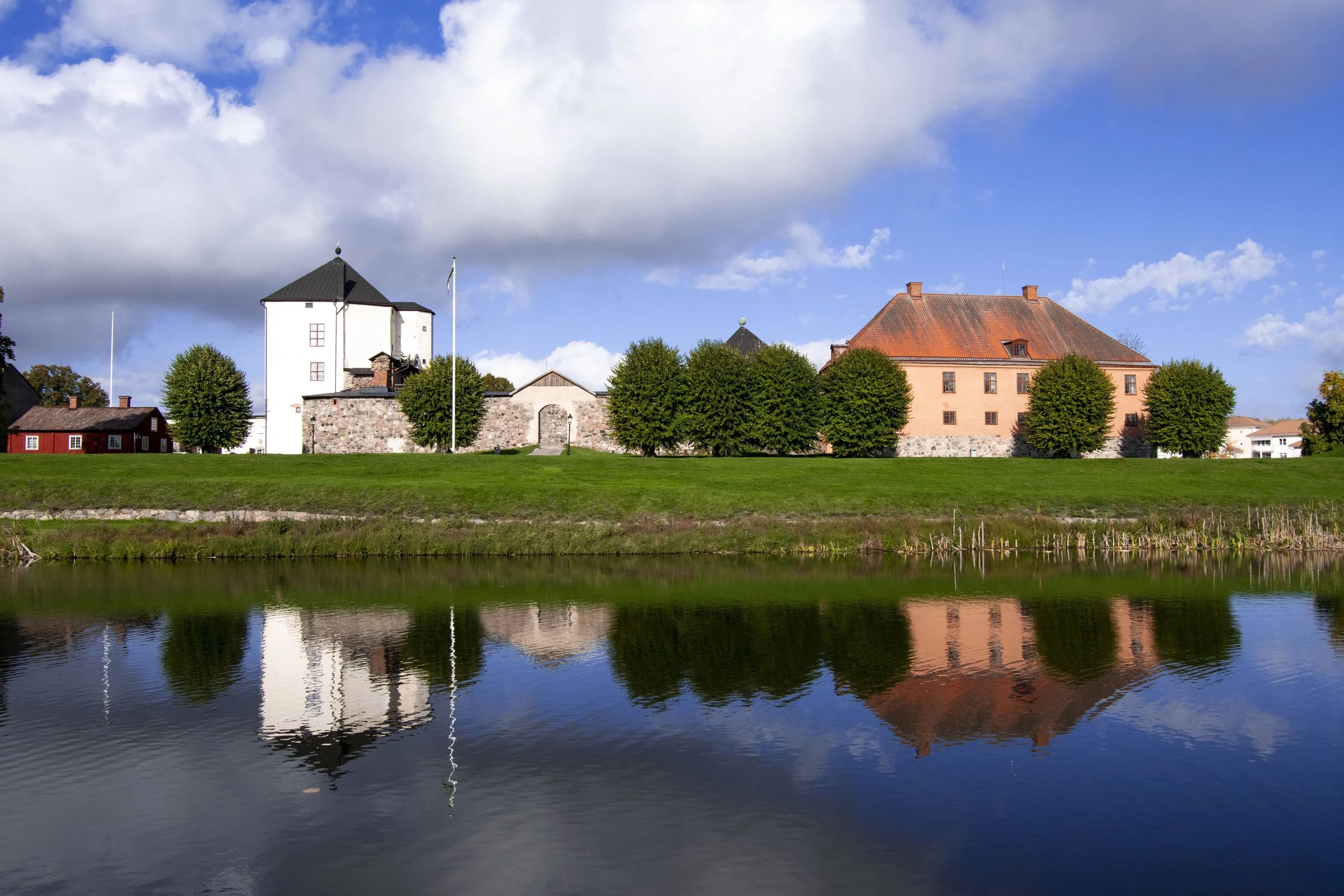 A scenic view of a river reflecting trees and historic buildings with a blue sky and scattered clouds.