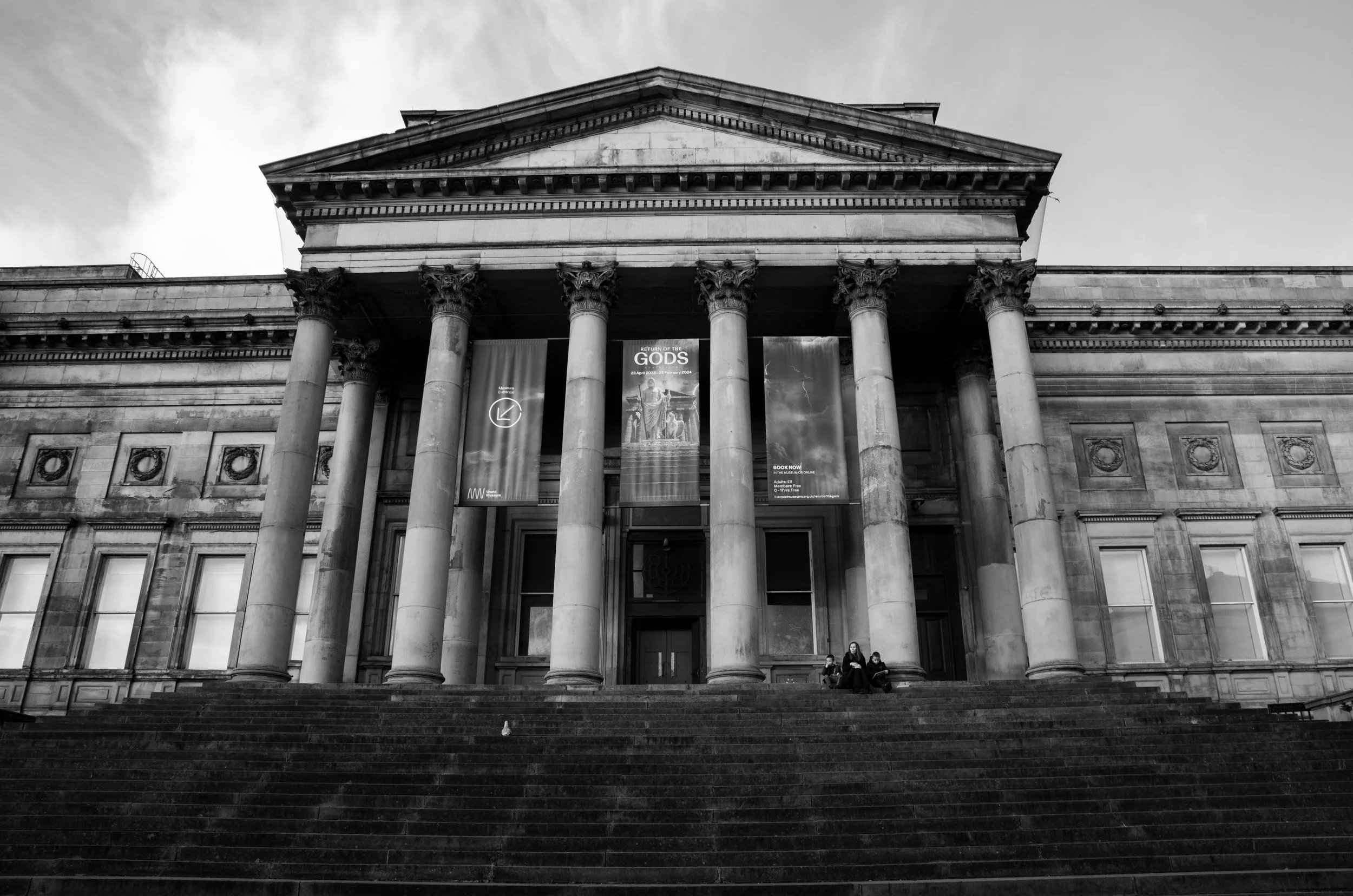 Black and white photo of a neoclassical building with tall columns and a triangular pediment. There are steps leading up to the entrance, where three people are sitting. Two banners advertising an exhibit titled 'Return of the Gods' hang between the columns.