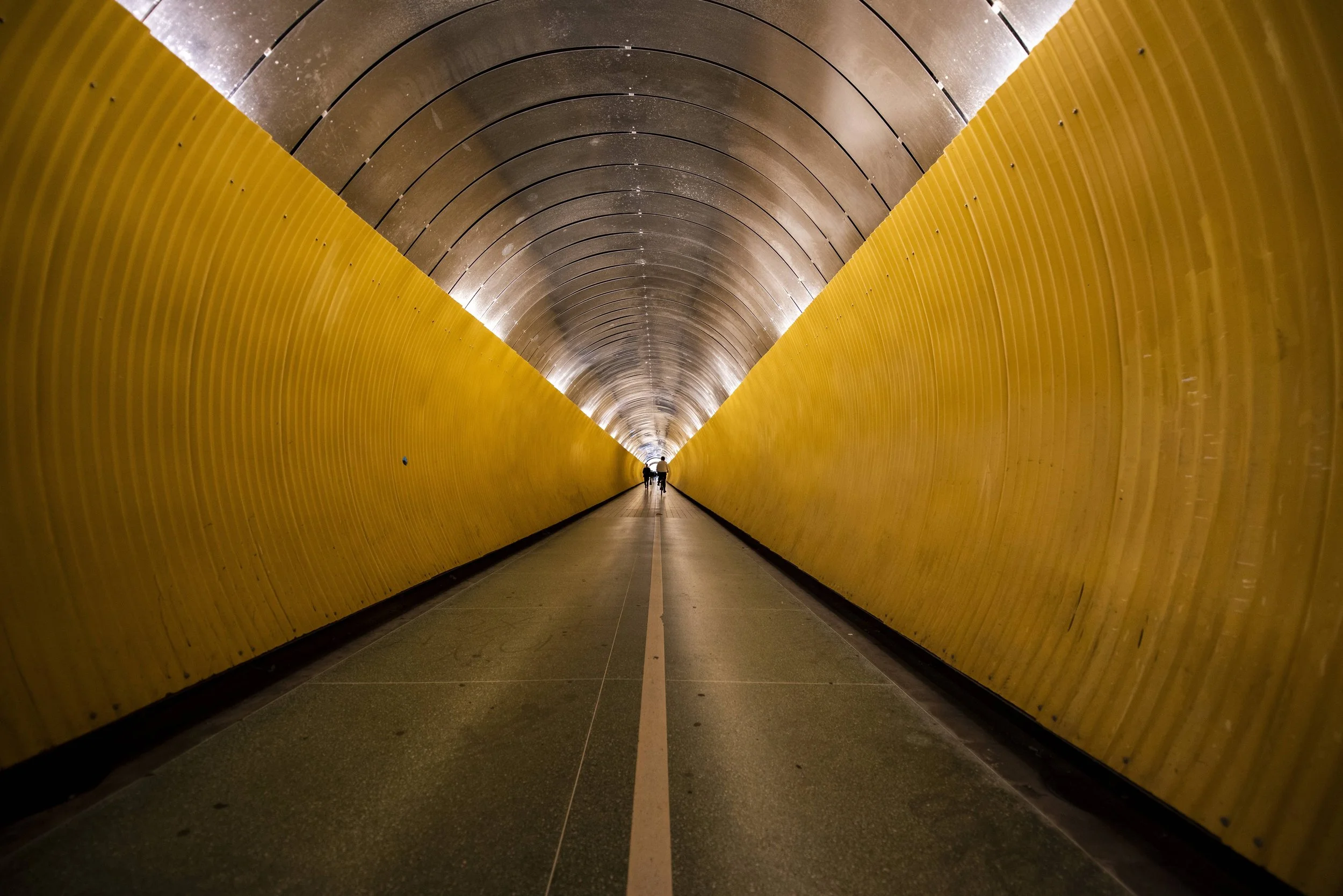 A long tunnel with a curved ceiling, featuring yellow walls on both sides. There are small lights illuminating the tunnel, and two people are walking in the distance towards the exit.