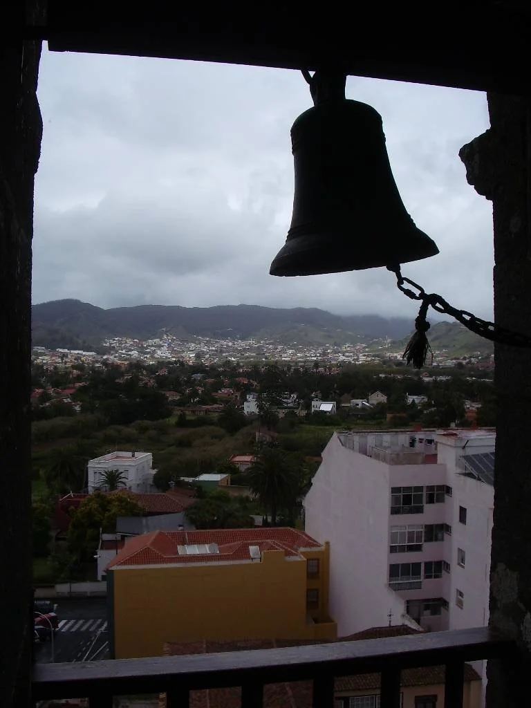 A large bell hanging from a wooden beam on a balcony, overlooking a city with mountains in the background. The sky is cloudy.