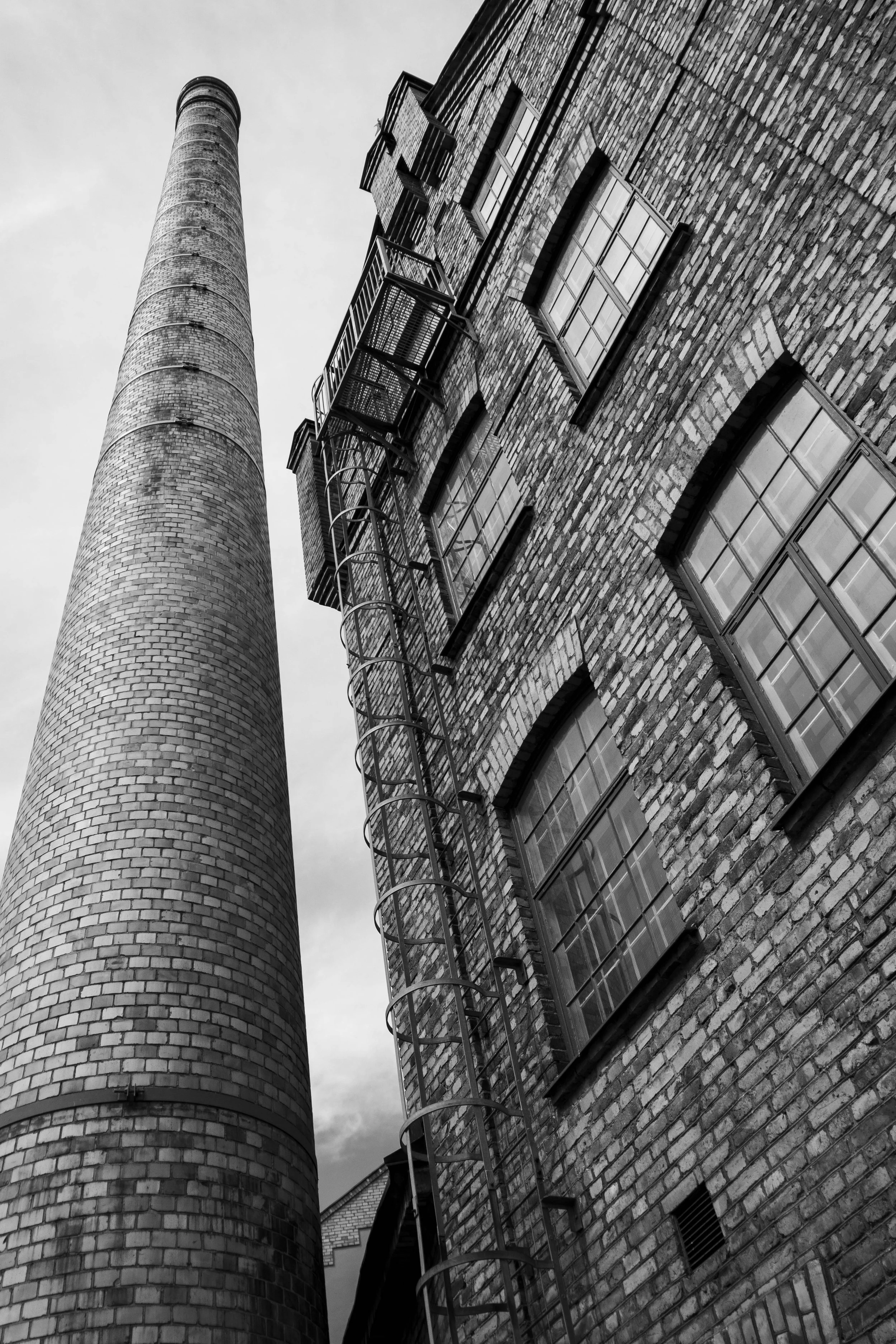 Black and white photo of a tall brick building with multiple windows and fire escapes, and a large brick smokestack in the foreground.
