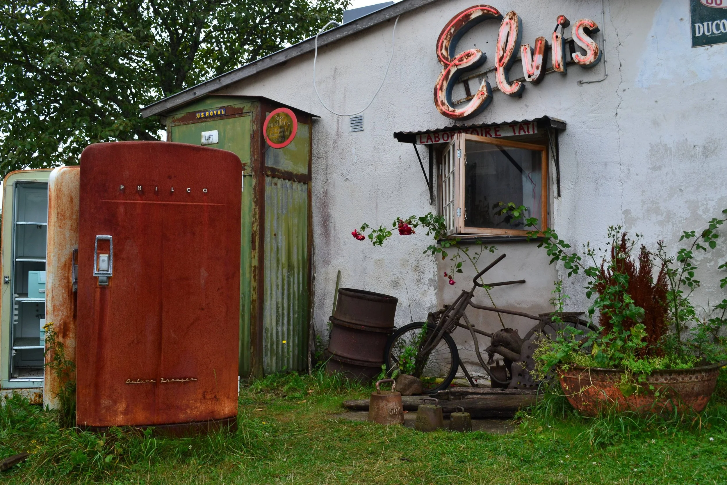 A vintage scene with an old red Philco refrigerator, weathered green metal structure, rusty bicycle, flowered plants, and worn sign on a building with a large neon "EUS" sign.