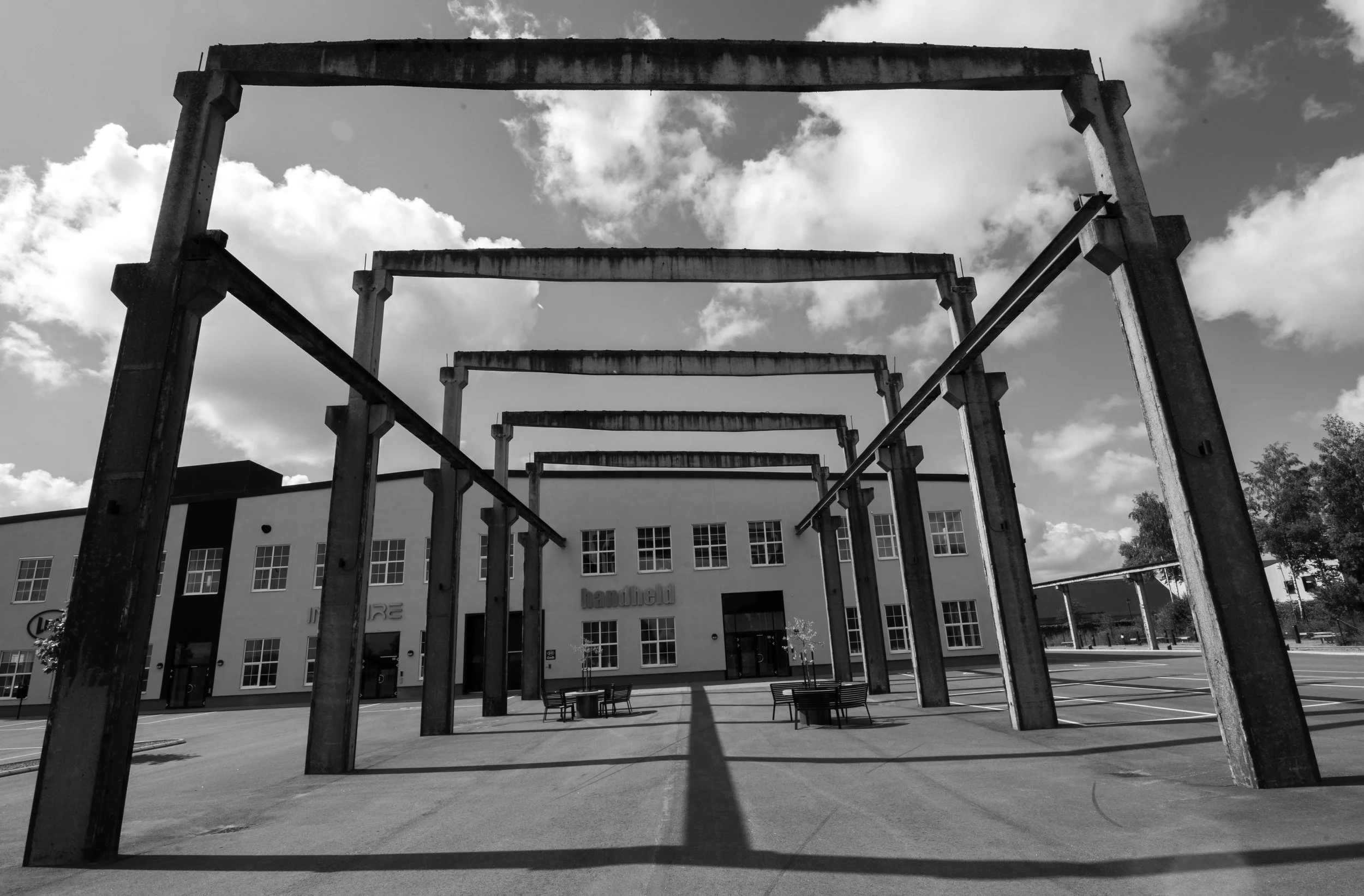 An outdoor schoolyard area with a large, modern building in the background, under a partly cloudy sky. In the foreground, there is a metal structure resembling a frame or scaffolding, with benches and a large open space underneath.