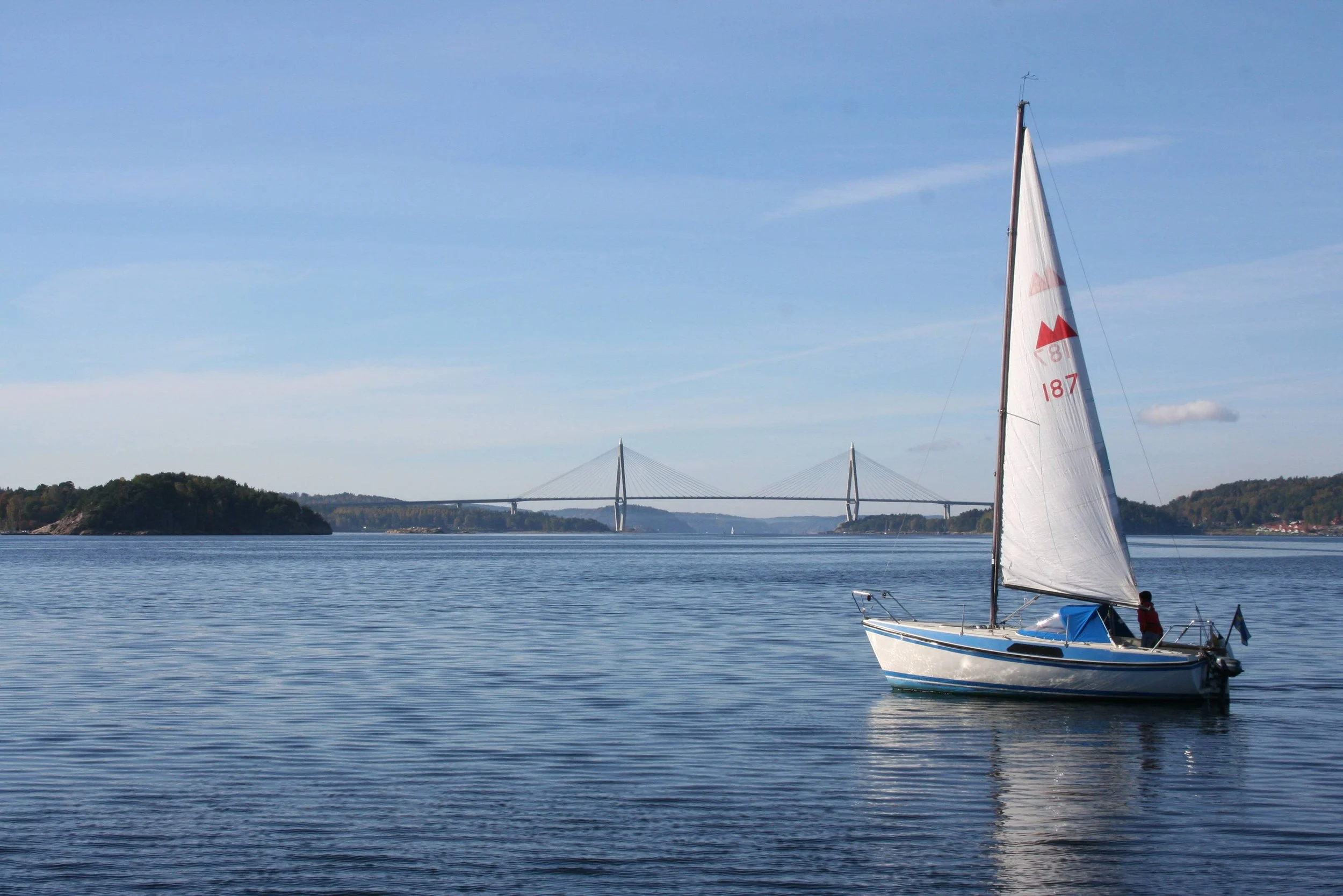 A sailboat with a white sail on calm water, a bridge in the background, and a small island with trees on the left under a clear blue sky.