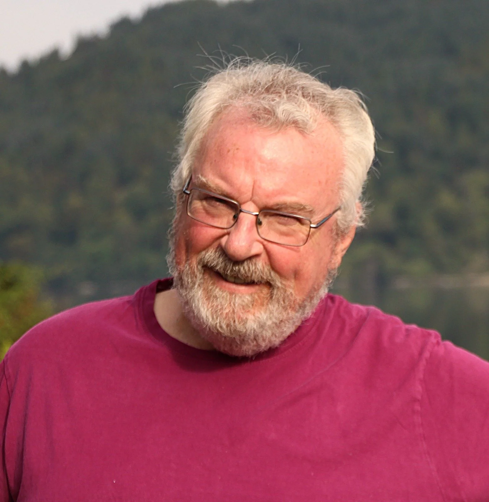 Smiling elderly man with glasses and a beard wearing a maroon shirt, outdoors with Trees in the background.
