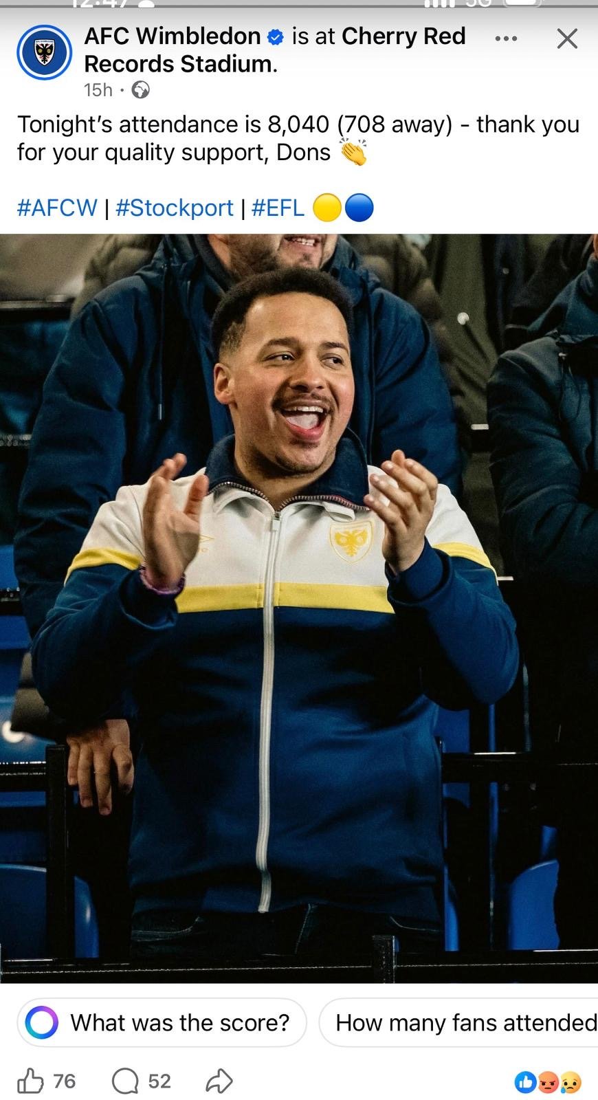 A man with short dark hair and a goatee clapping and smiling while attending a sporting event at Cherry Red Records Stadium. He is wearing a sports jacket with a crest on the chest, and other spectators are visible around him.