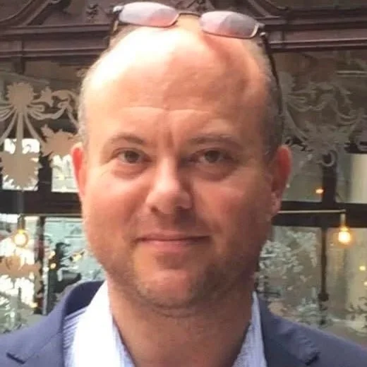 Close-up of a man with glasses resting on his forehead, wearing a dark blazer over a light shirt, outside in front of a decorative glass window.