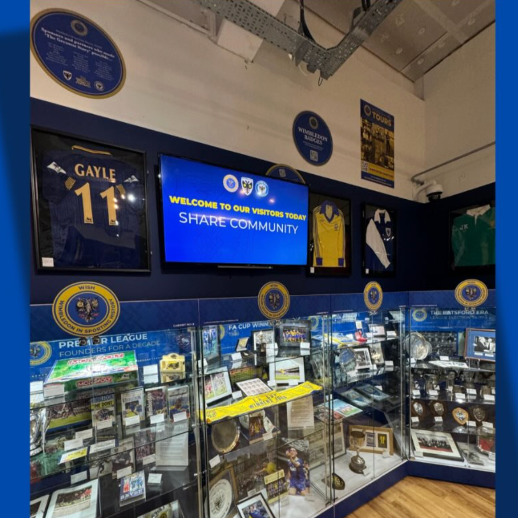 A display case with AFC Wimbledon sports memorabilia at a sports museum, featuring framed shirts, badges, trophies, and digital screens with informational signs and images.