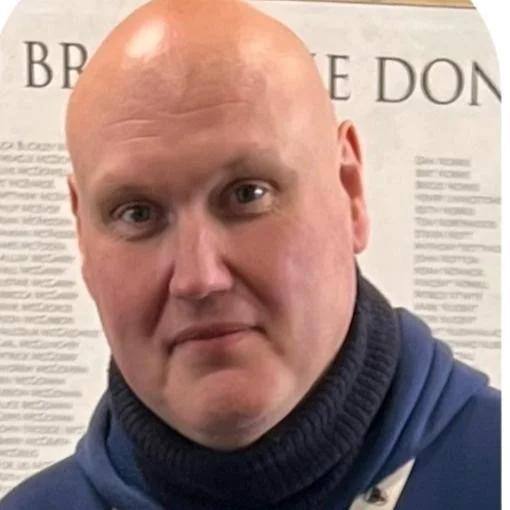 A bald man wearing a dark blue hoodie stands in front of a wall with text, looking at the camera.