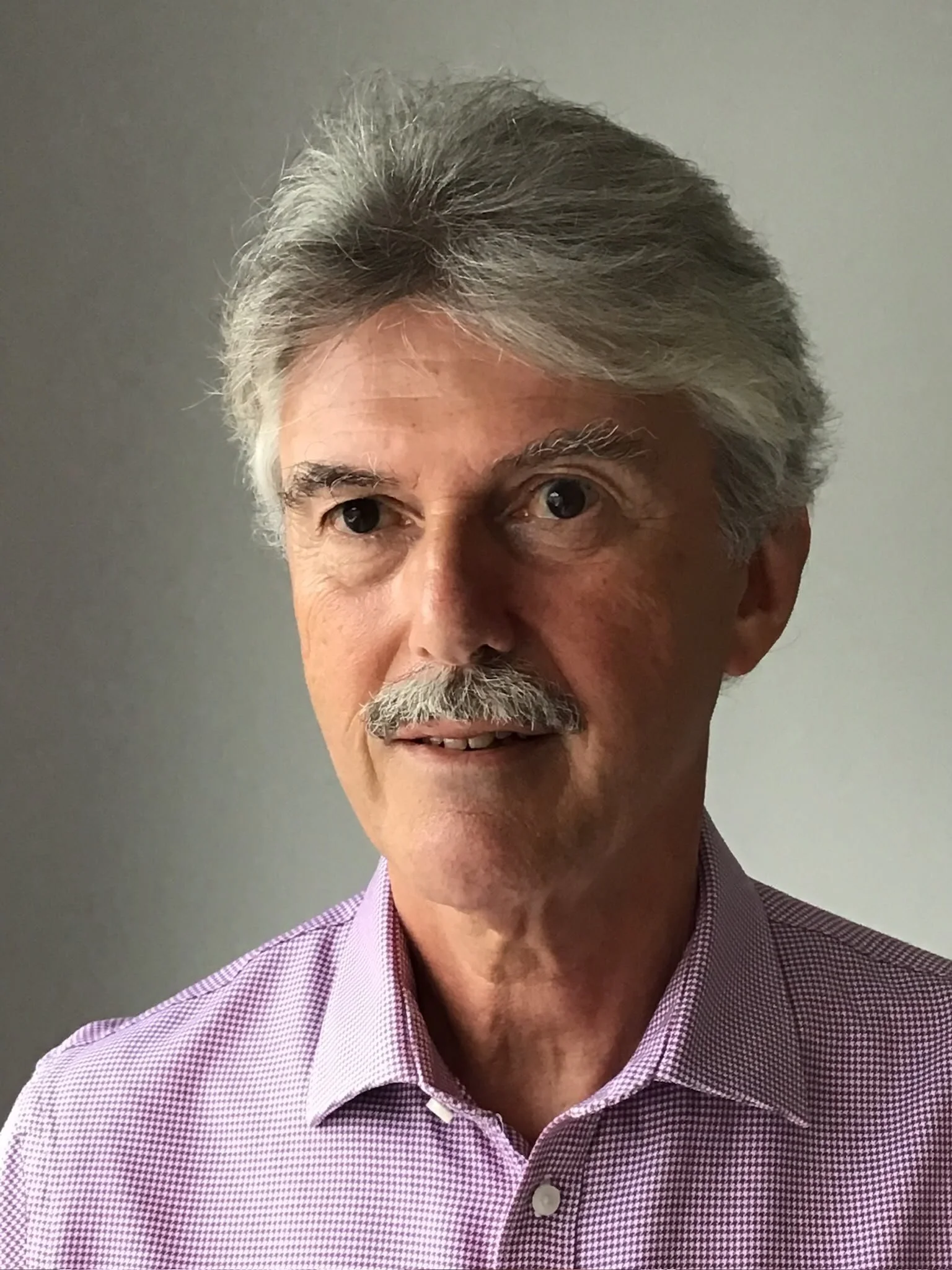 Close-up of a middle-aged man with gray hair, mustache, and beard, wearing a purple checkered shirt, smiling slightly, against a plain gray background.