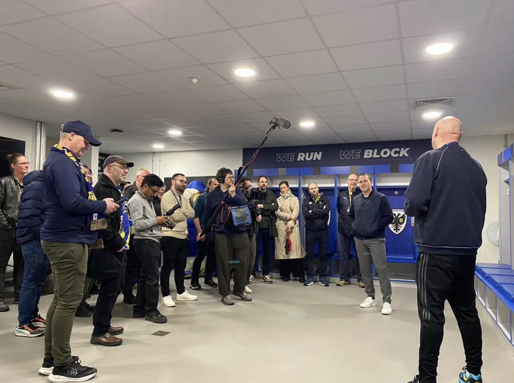 A group of people in a changing room, some wearing sports hoodies and scarves, gathered around a man running a stadium tour. a camera crew is recording the scene with a boom microphone.
