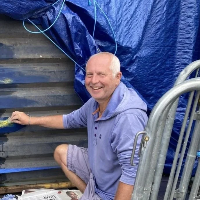 A smiling man in a light purple hoodie and shorts, kneeling beside a pond or water feature, with a blue tarp and blue ropes in the background.