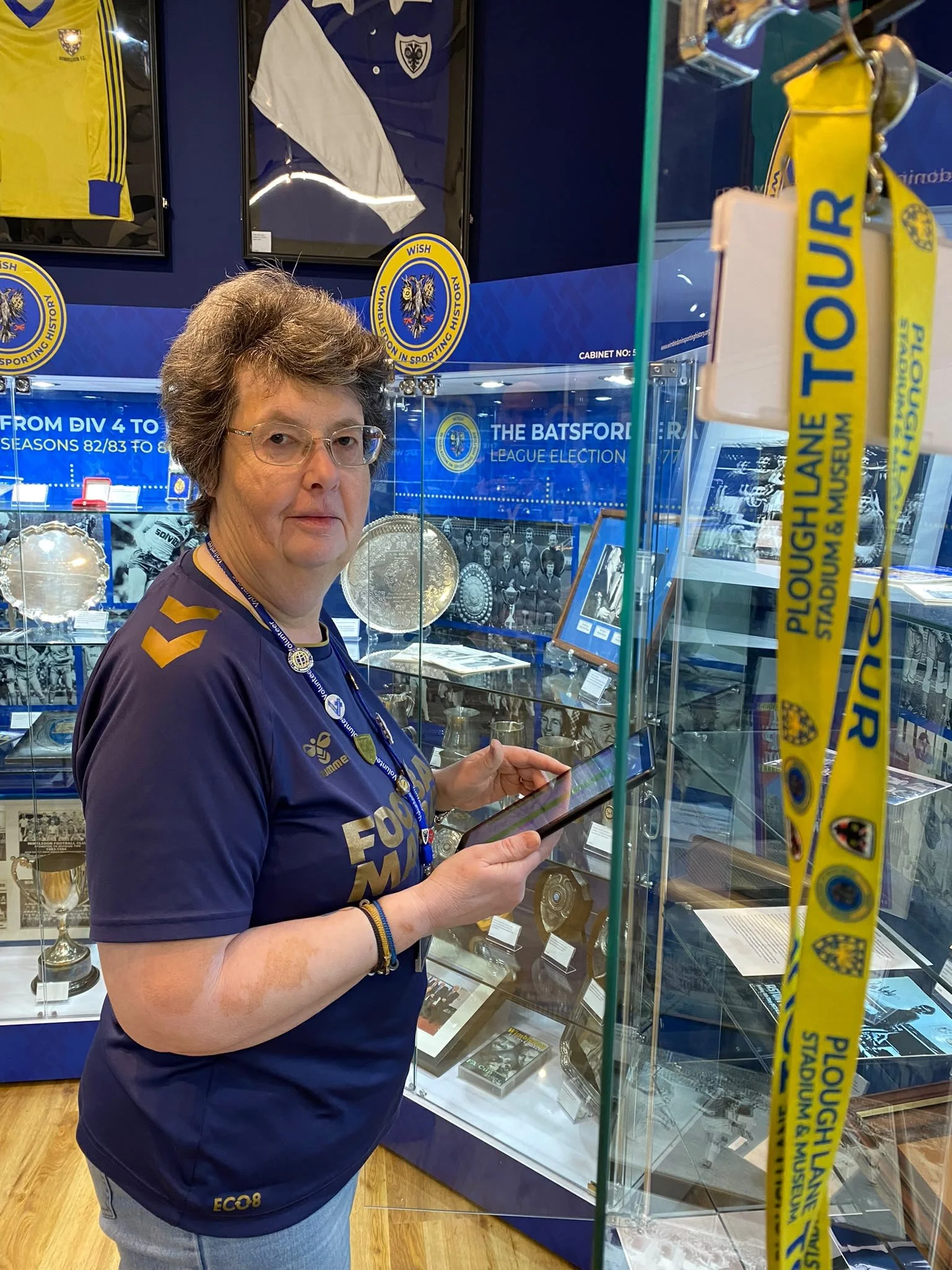 A woman with glasses and short, curly hair, the museum curator, holding a tablet in a display case with sports memorabilia, including framed football shirts and silverware, at a museum dedicated to AFC Wimbledon football history.