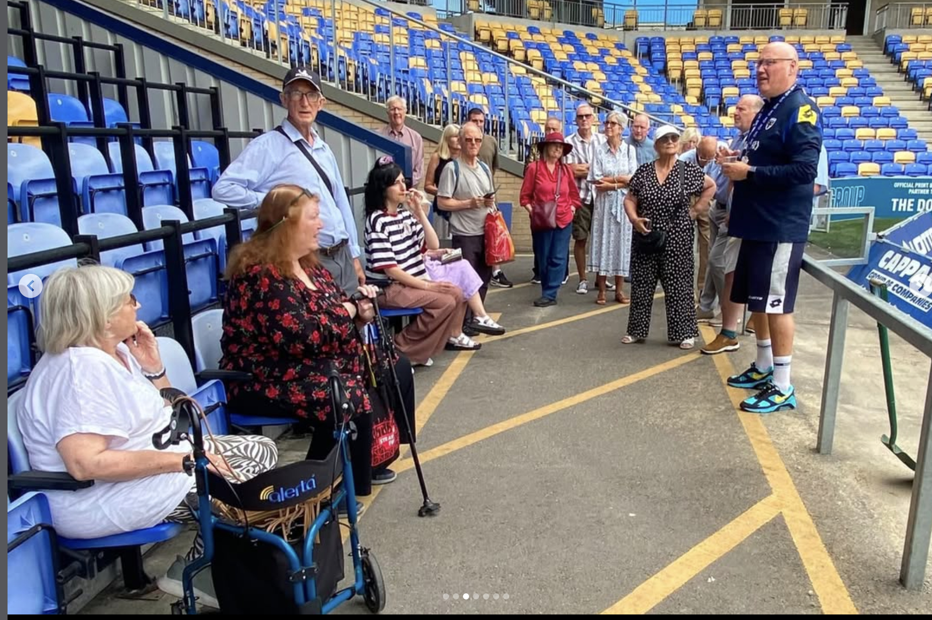 A group of people gathered at at afc wimbledon stadium tour, some sitting in blue seats and others standing, listening to a man in a blue jacket giving a speech or presentation near the stand